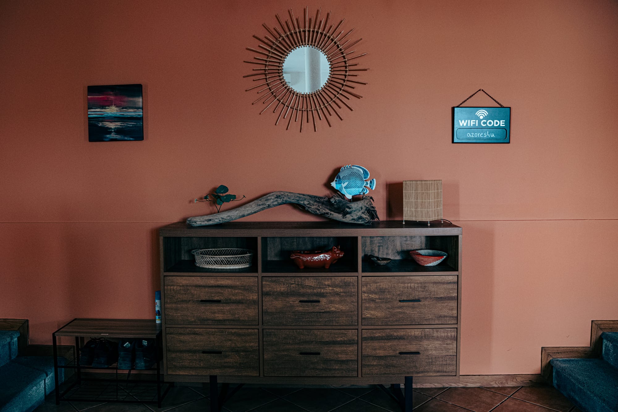 A warmly lit entryway at Lava Coliving featuring a sunburst mirror, orange wall, wooden dresser with decorative items, and a sign displaying the wifi code