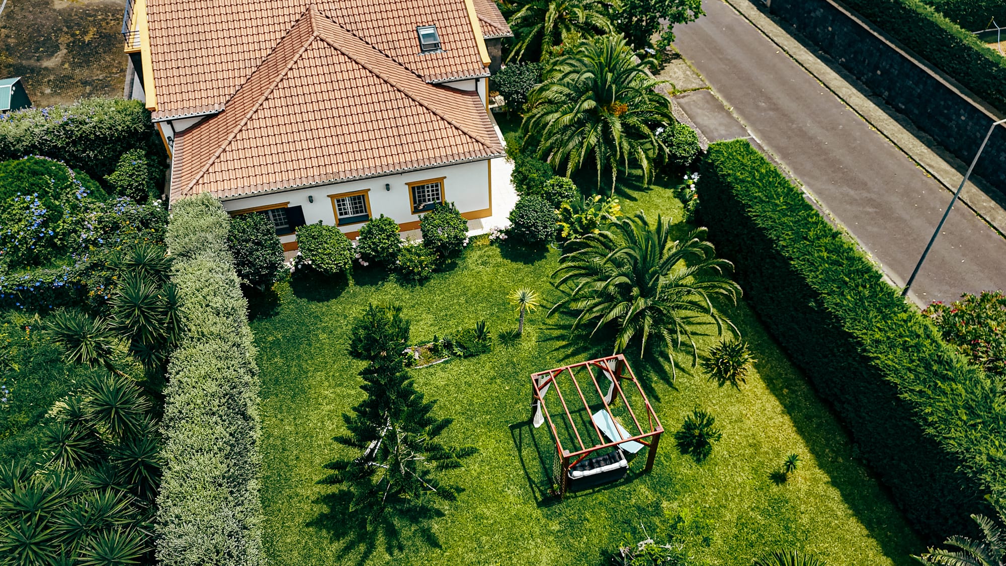 Aerial view of Lava Coliving showing a large green garden with palm trees, hedges, a red wooden swing frame, and part of the white and yellow house with a tiled roof