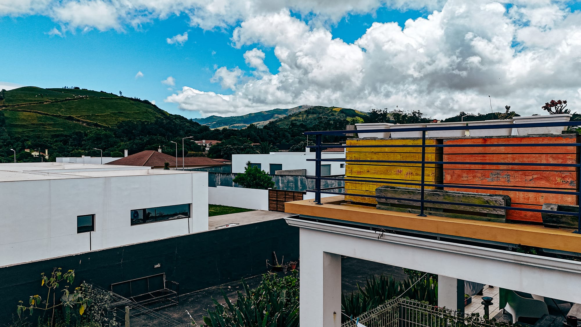 Colorful wooden bench and railing on a balcony at Lava Coliving, overlooking green hills, modern white buildings, and a blue sky filled with puffy clouds