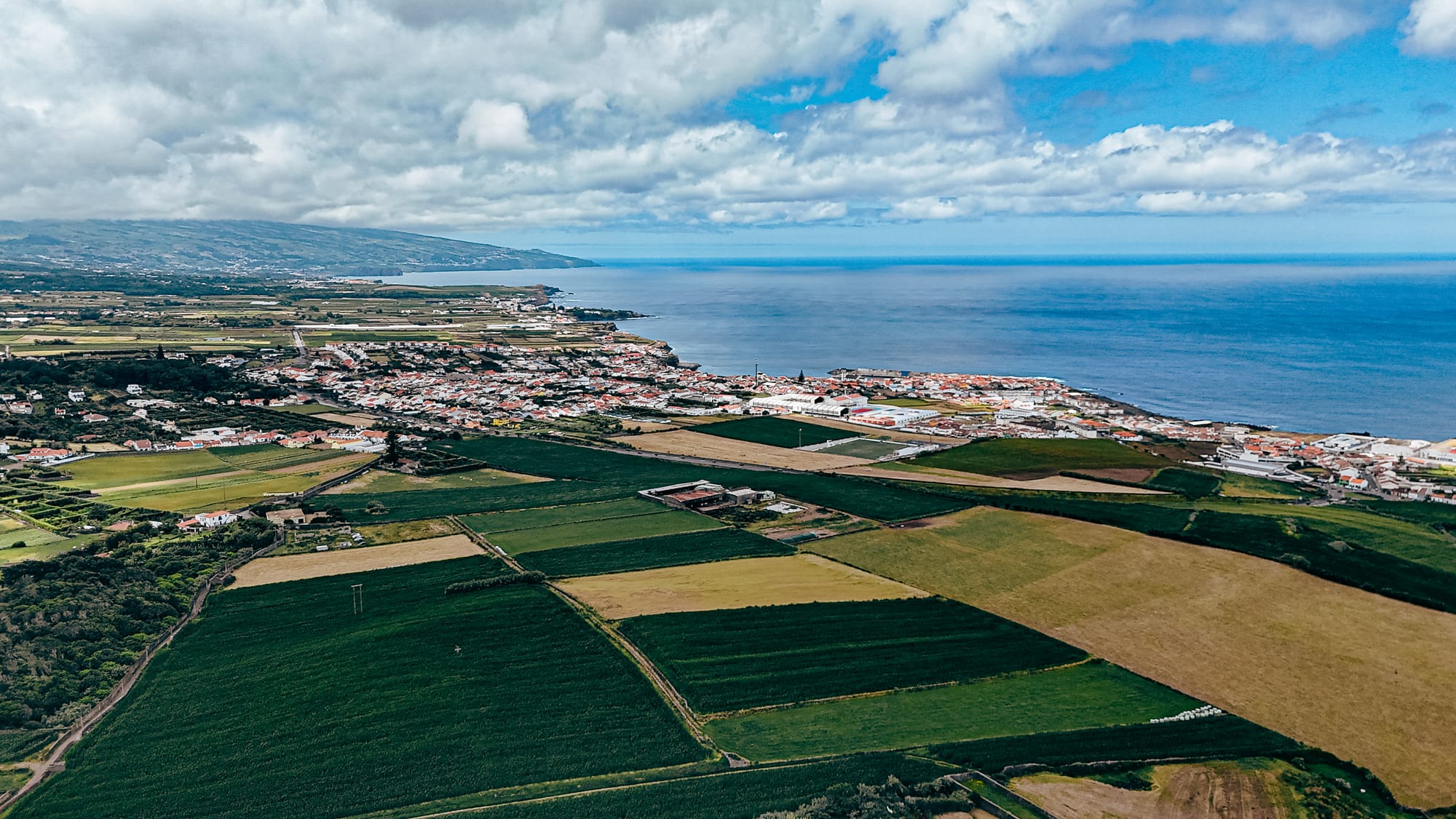 Aerial view of the coastline near Lava Coliving, showing patchworked green farmland, a coastal town with red-roofed buildings, and the deep blue Atlantic Ocean under a partly cloudy sky