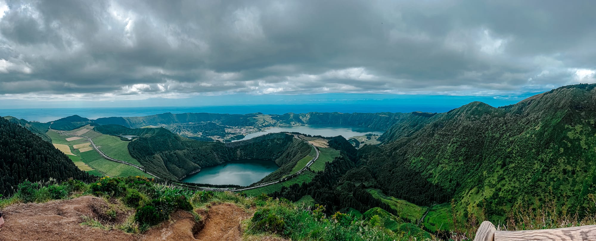 Wide-angle view from Miradouro da Grota do Inferno showing twin crater lakes, winding roads, and rugged volcanic ridgelines beneath a brooding sky