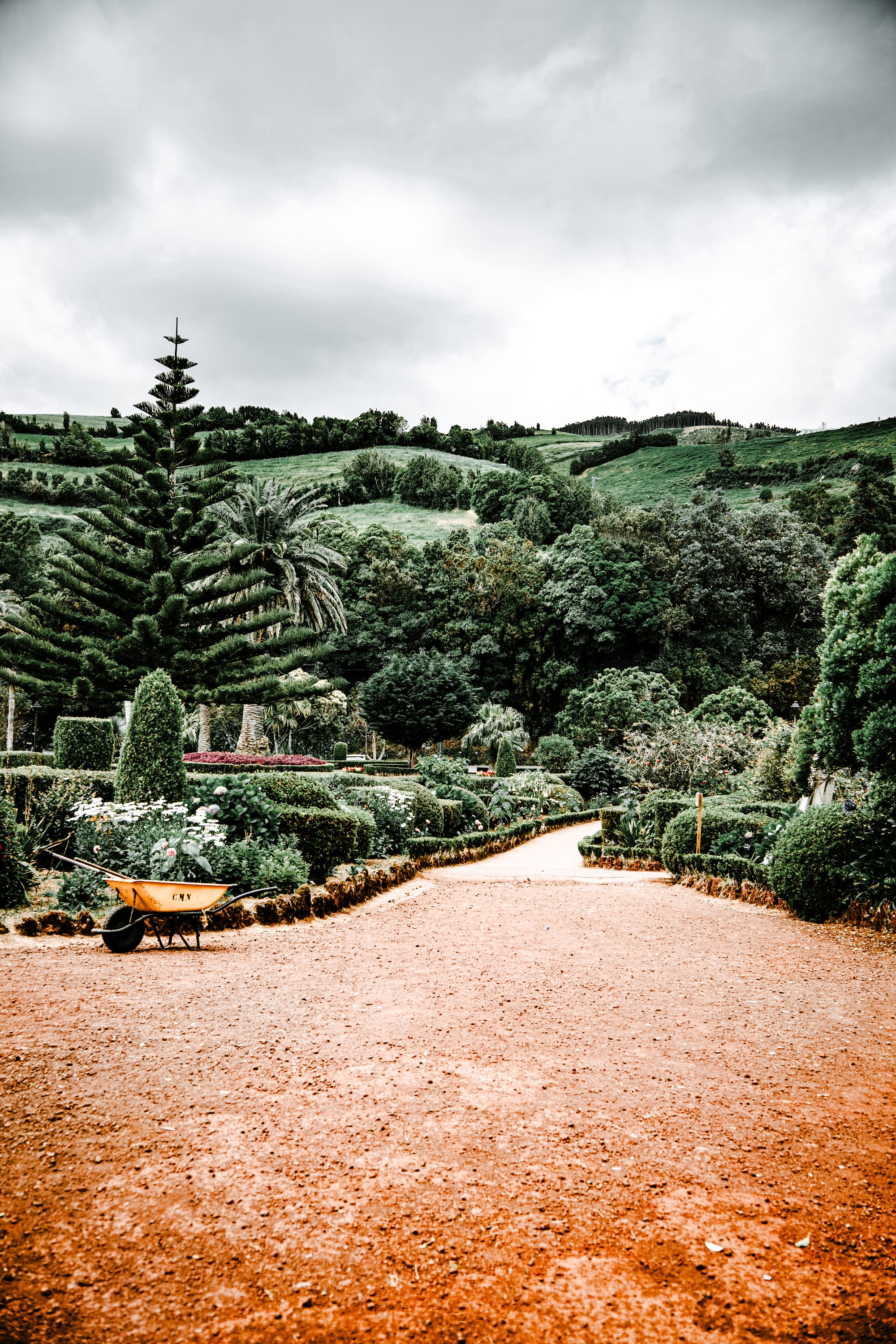 Pathway through lush gardens with manicured hedges and a yellow wheelbarrow, backed by forested hills