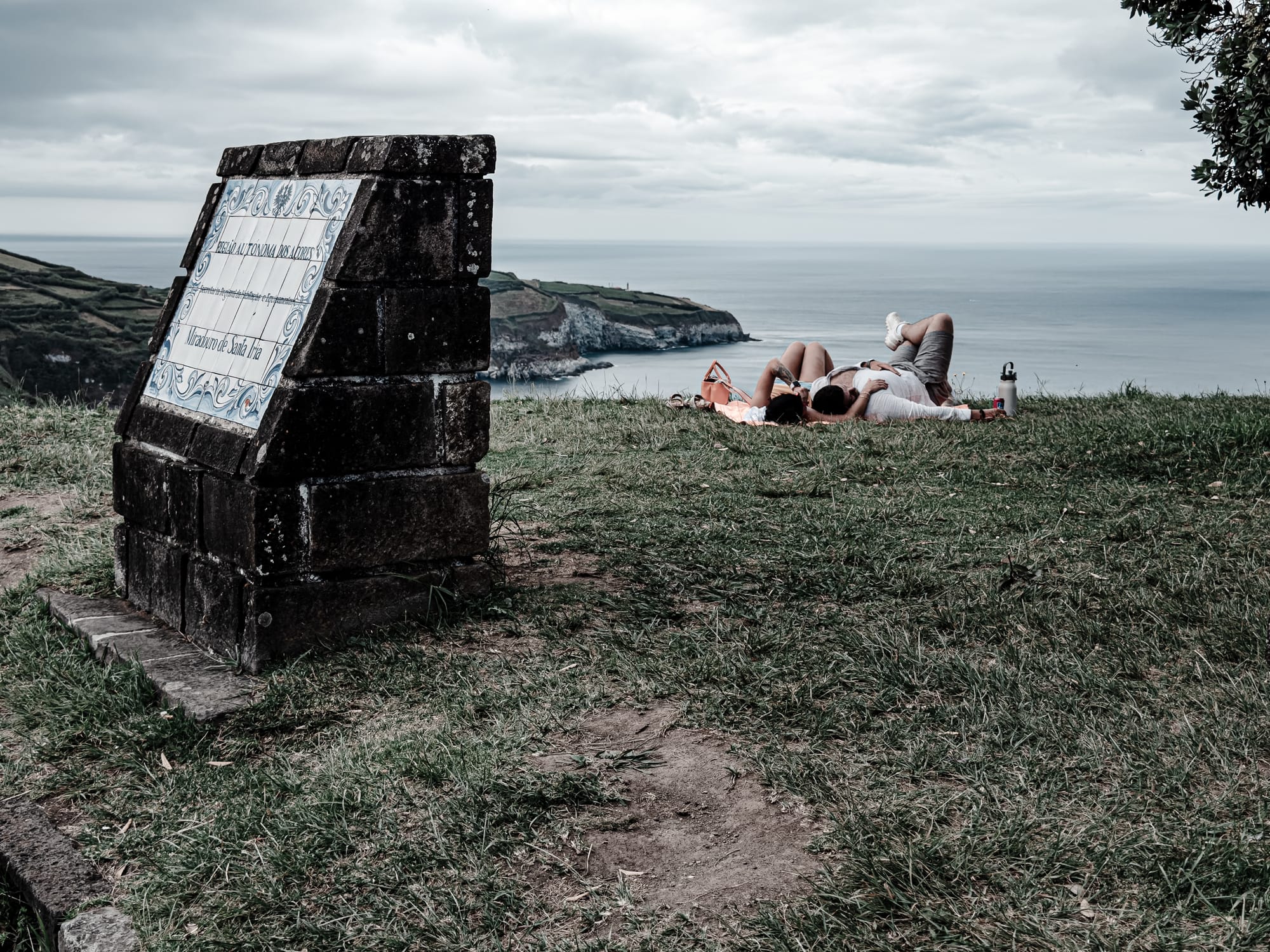 Couple lying on a grassy overlook near the Miradouro de Santa Iria sign, with cliffs and ocean in the background