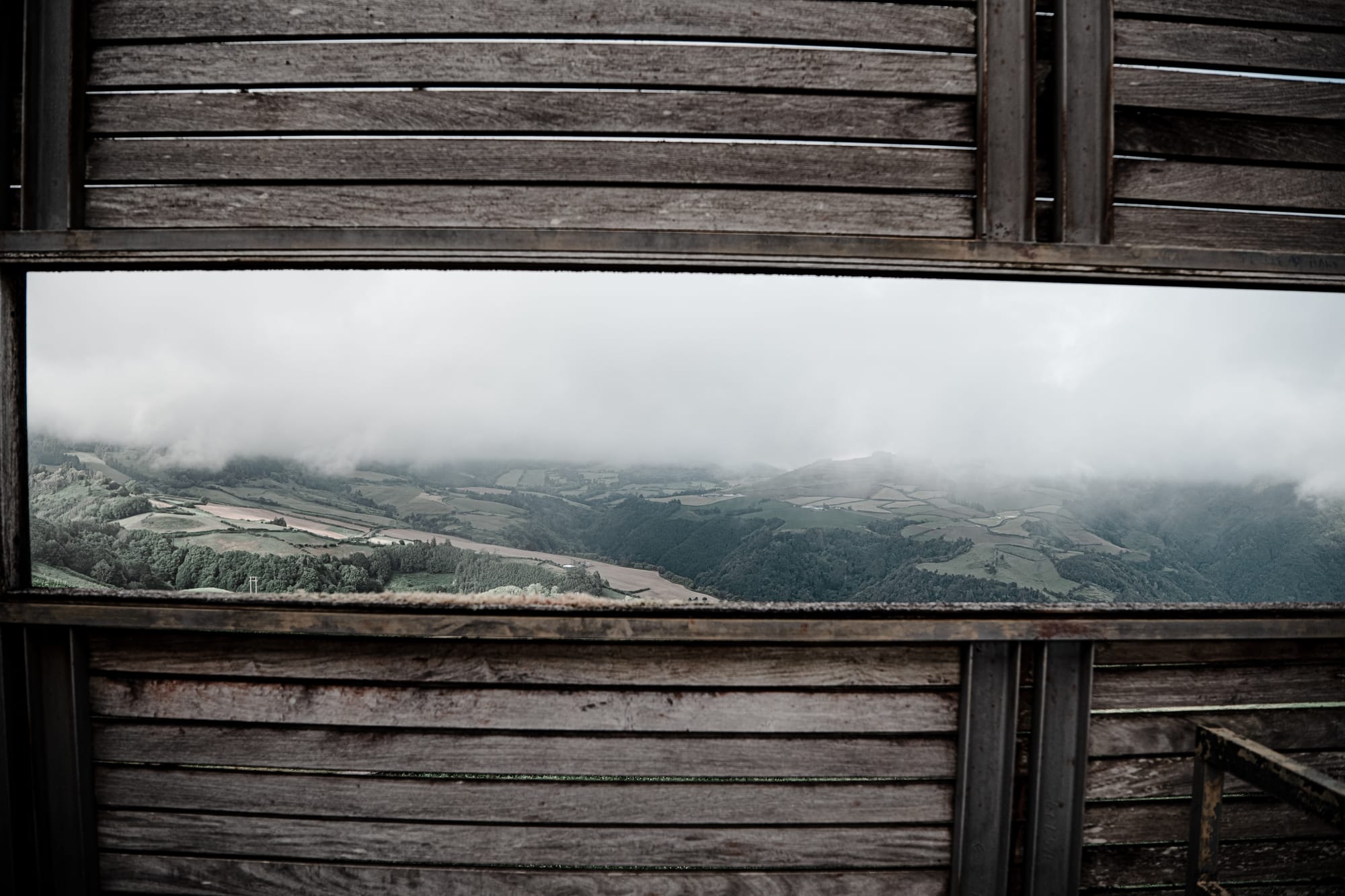 View through a rectangular wooden window frame overlooking misty green hills and farmland