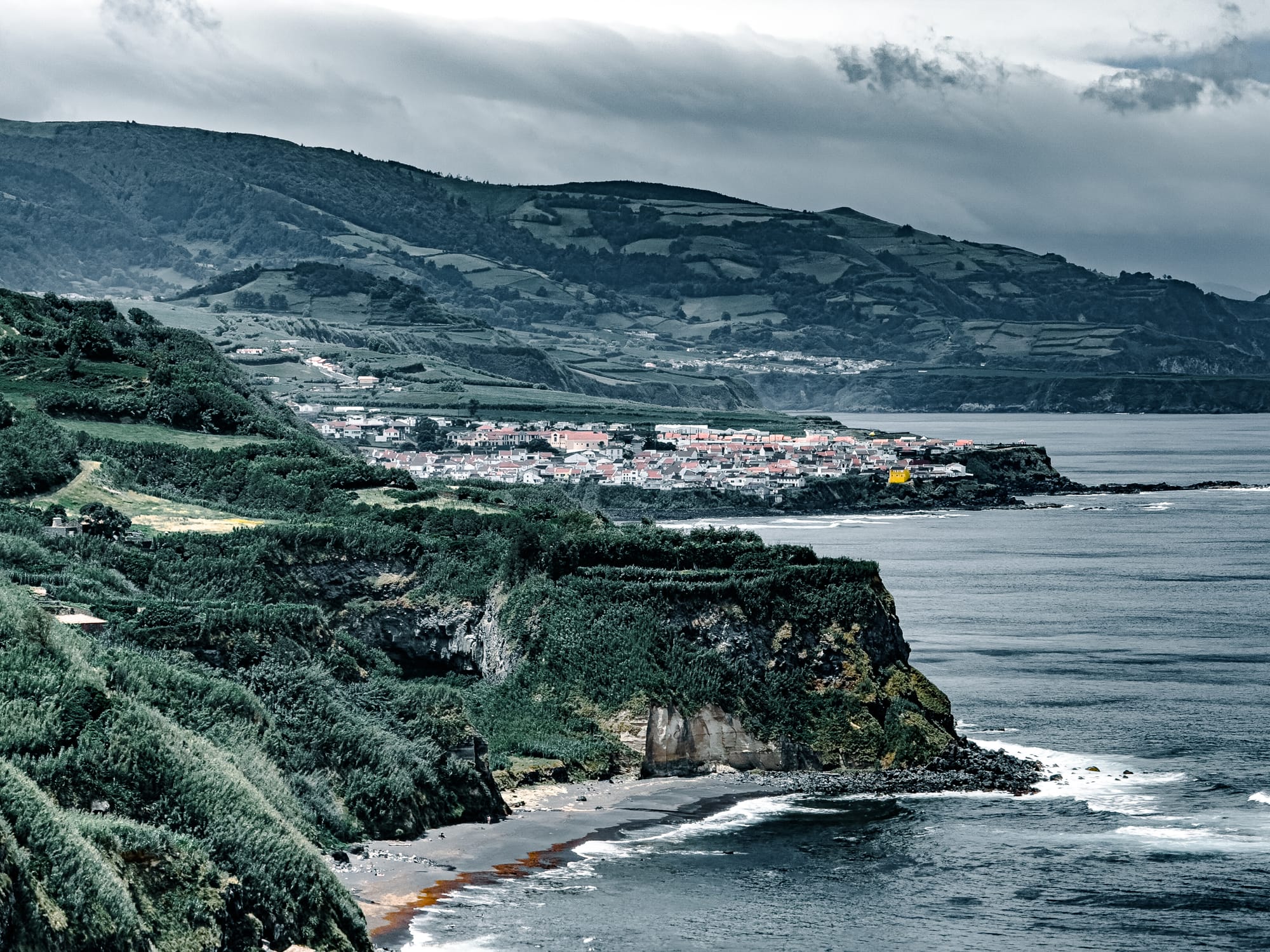 A coastal town sits at the edge of the sea, framed by dramatic green cliffs and terraced hills, with dark sand beaches and waves breaking below