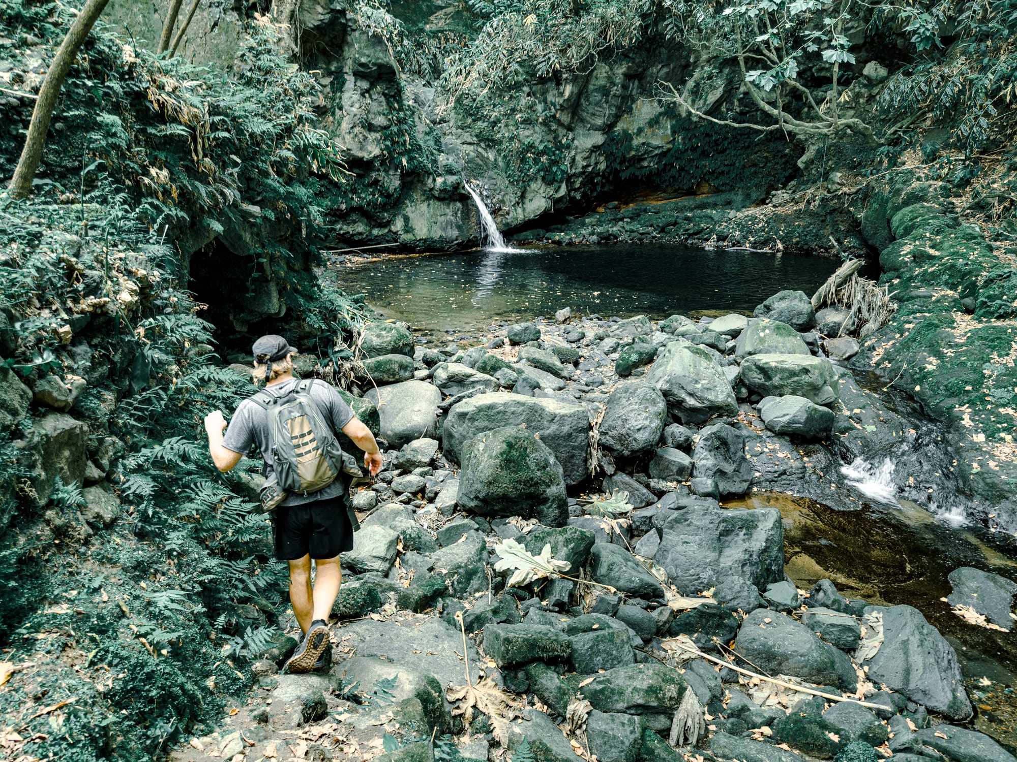Hiker walking across volcanic rocks toward a small waterfall cascading into a dark forest pool