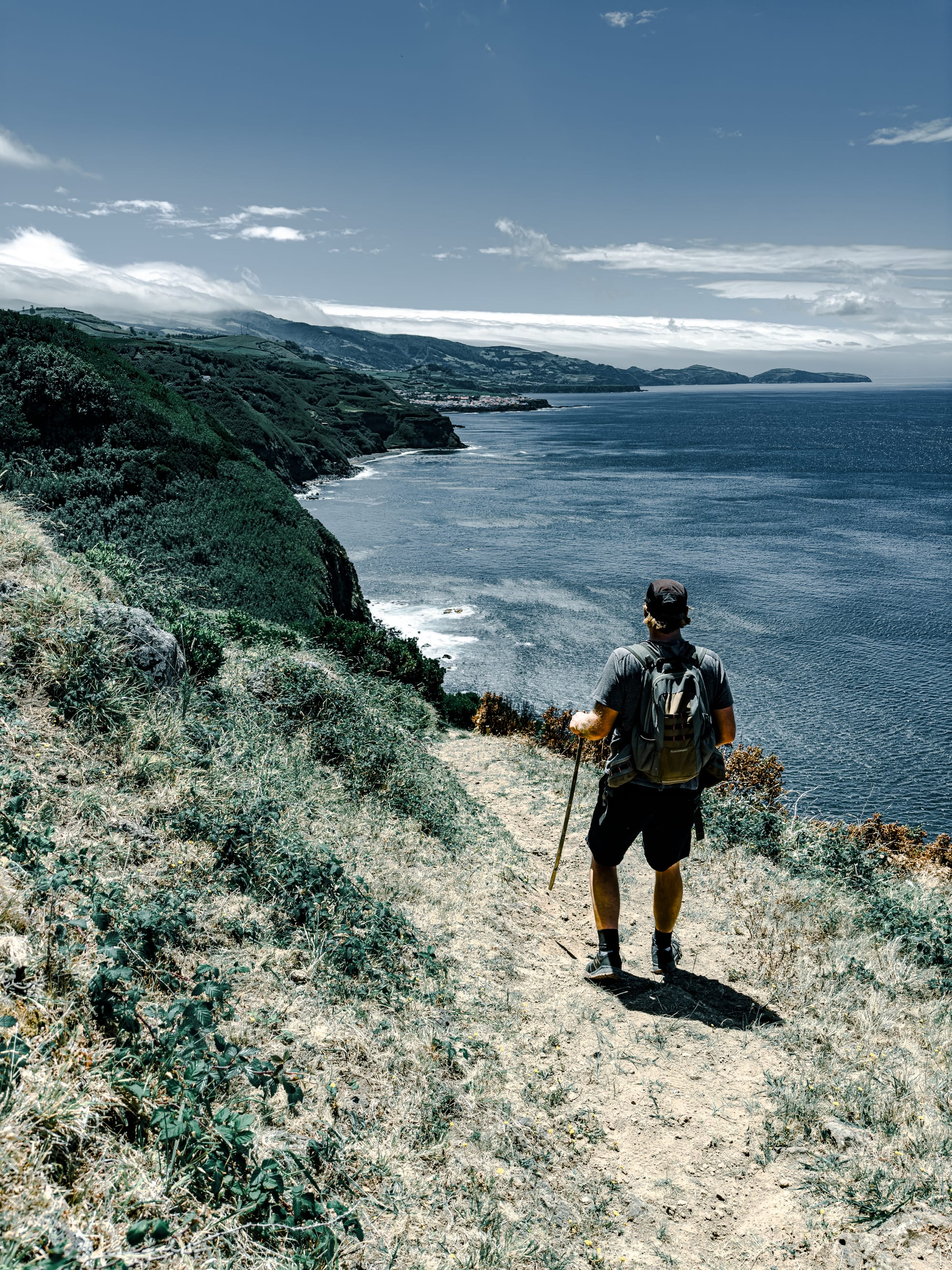 Guide Chris with a backpack and walking stick stands on a dirt trail overlooking steep green cliffs and the Atlantic Ocean, with a coastal town visible in the distance