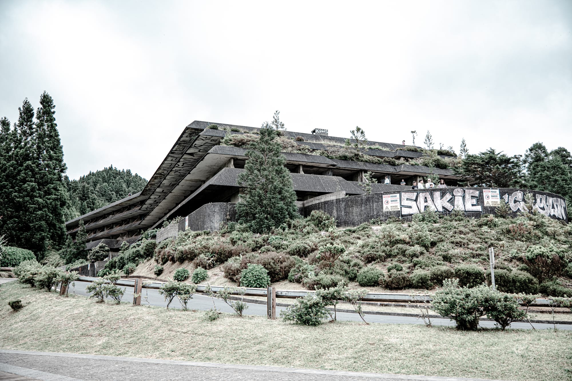 Wide-angle view of the abandoned Monte Palace Hotel in São Miguel, Azores, showing its angular, terraced architecture partially overtaken by moss and greenery, with graffiti across its lower walls and tall evergreens surrounding it