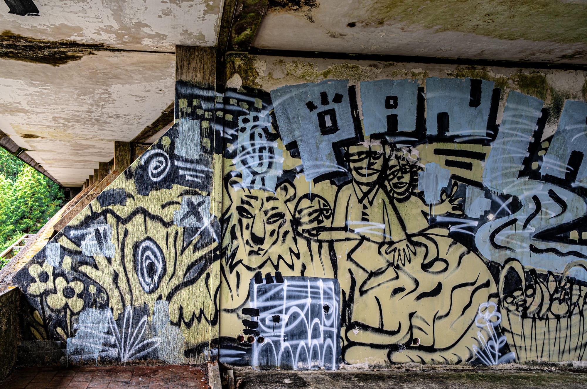 Graffiti mural in an abandoned hotel featuring stylized black-line figures including a smiling couple, a tiger, and surreal plant-like elements on a faded yellow background with overlapping tags and white spray paint marks