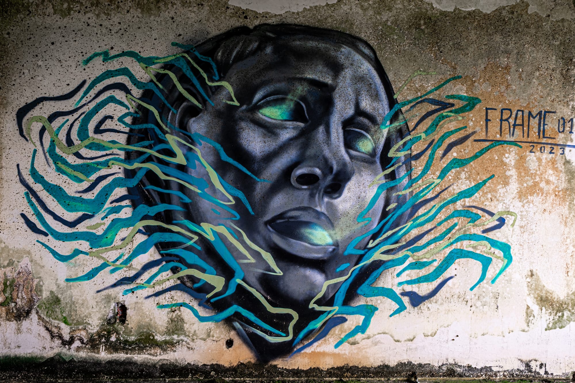 Graffiti mural of a dark-toned face with glowing green eyes and lips, surrounded by swirling teal and blue lines, signed FRAME01 on a weathered concrete wall