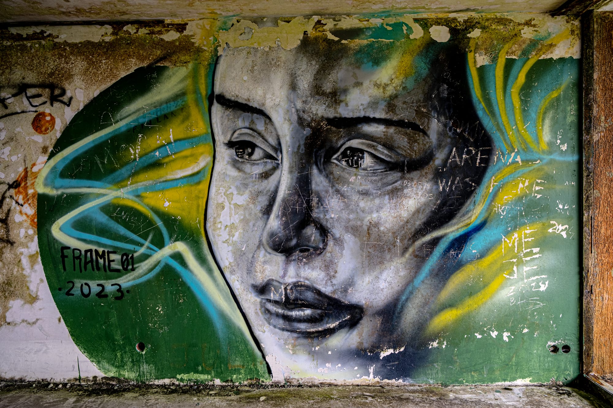 Graffiti mural of a woman’s face with green and yellow abstract lines, painted inside an abandoned hotel room with peeling walls and etched graffiti marks