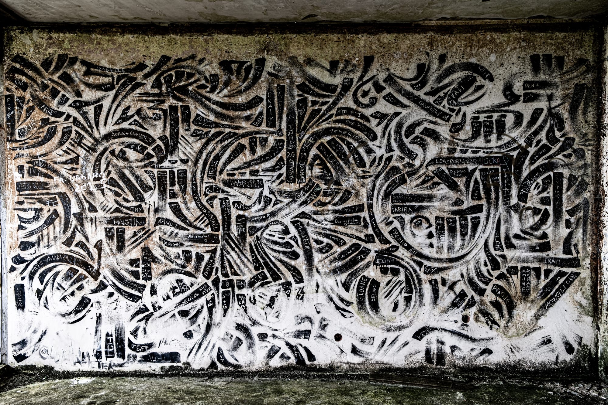 Abstract black and white mural composed of curving lines and dense patterns, covered in scratched-in names and dates on a weathered wall inside the abandoned Monte Palace Hotel