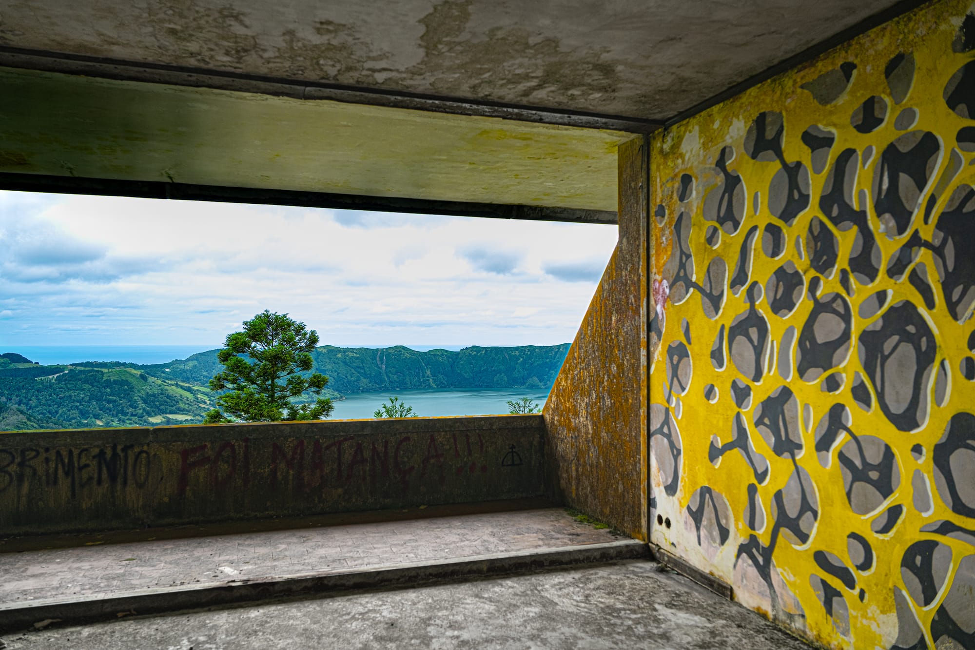 Graffiti-covered wall with abstract yellow and black mural inside an abandoned hotel room, with a window view of the Sete Cidades lakes and green mountains in the distance