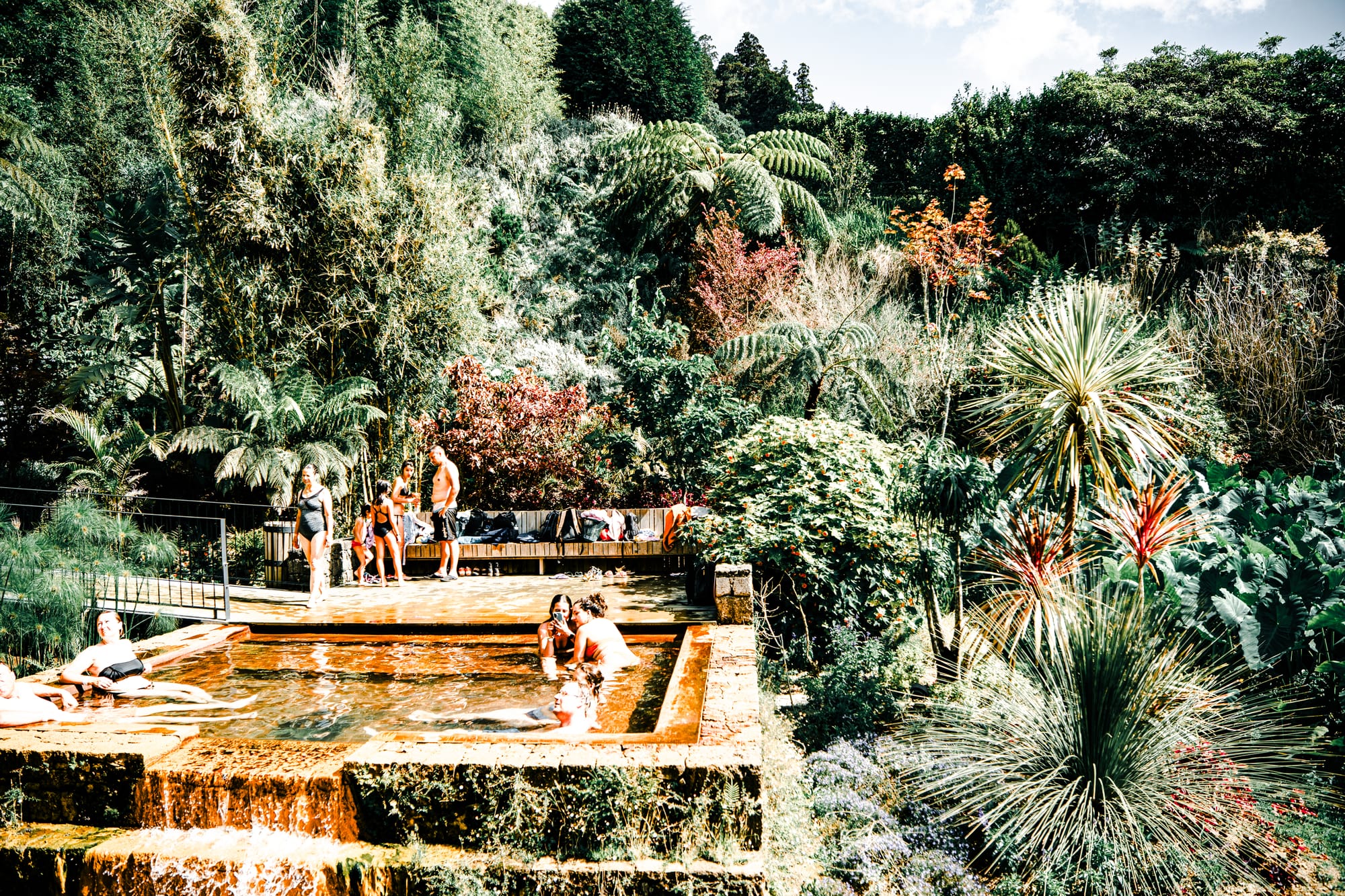 Visitors relaxing in a terraced thermal pool at Poça da Dona Beija, framed by dense tropical greenery and volcanic stone