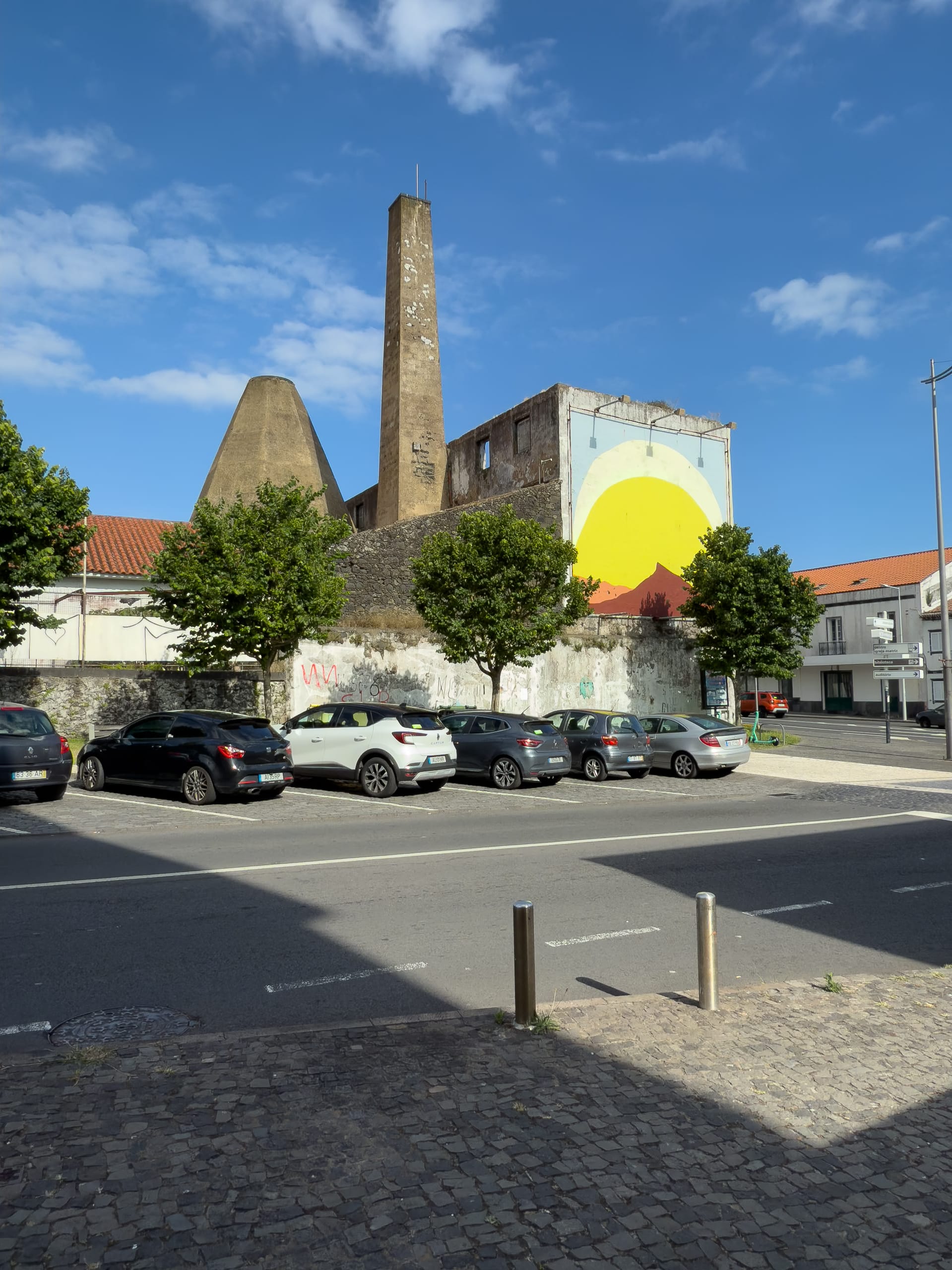 A bold yellow sun mural painted on the side of a weathered industrial building with a tall chimney, framed by small trees and a parking lot in Ponta Delgada