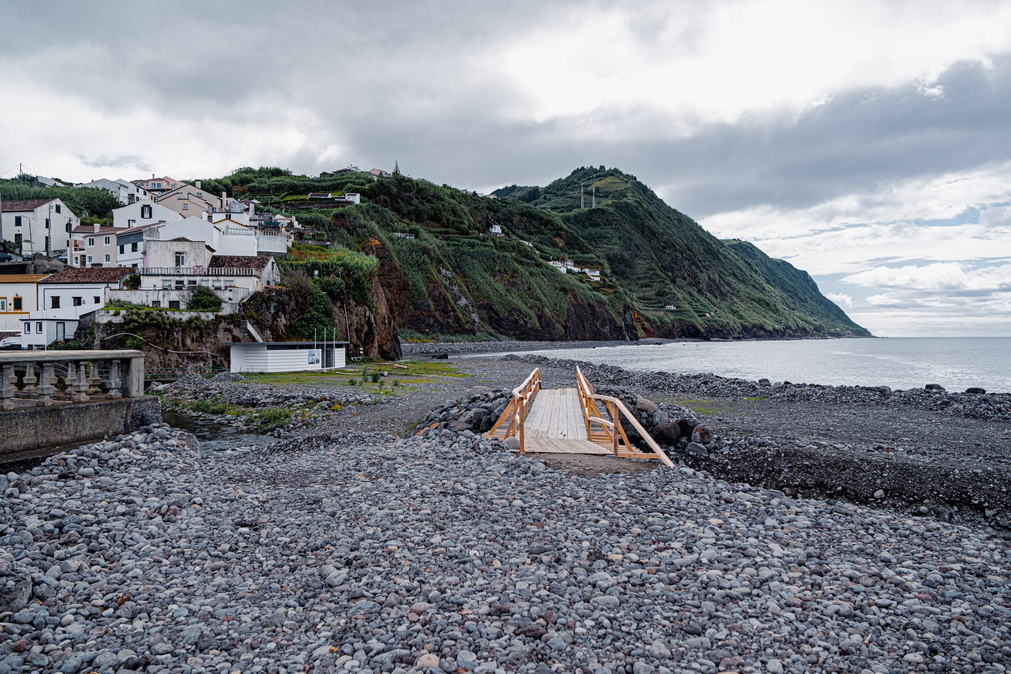 View of a small wooden bridge leading over rounded basalt stones toward the ocean in Povoação, São Miguel, with white hillside houses and steep green cliffs in the background under a cloudy sky
