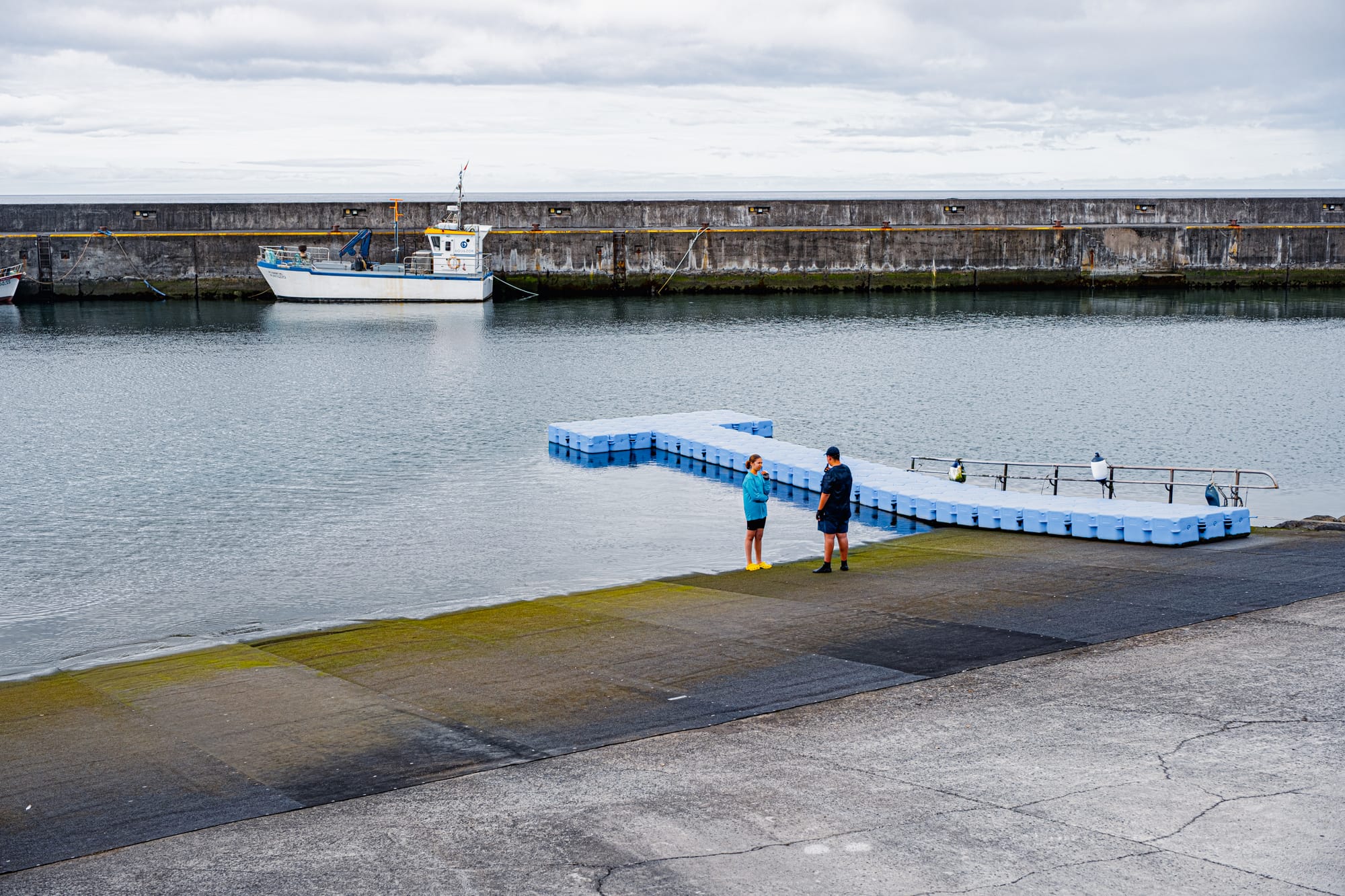 Two people stand near a blue floating dock in Povoação’s marina with a fishing boat moored in the background and concrete ramp leading into calm harbor water