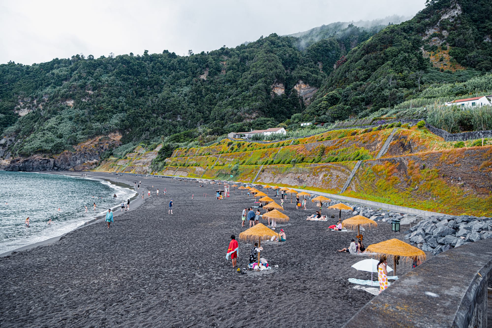 People relaxing under straw umbrellas on Praia do Fogo, Ribeira Quente, with dark volcanic sand and steep green cliffs in the background