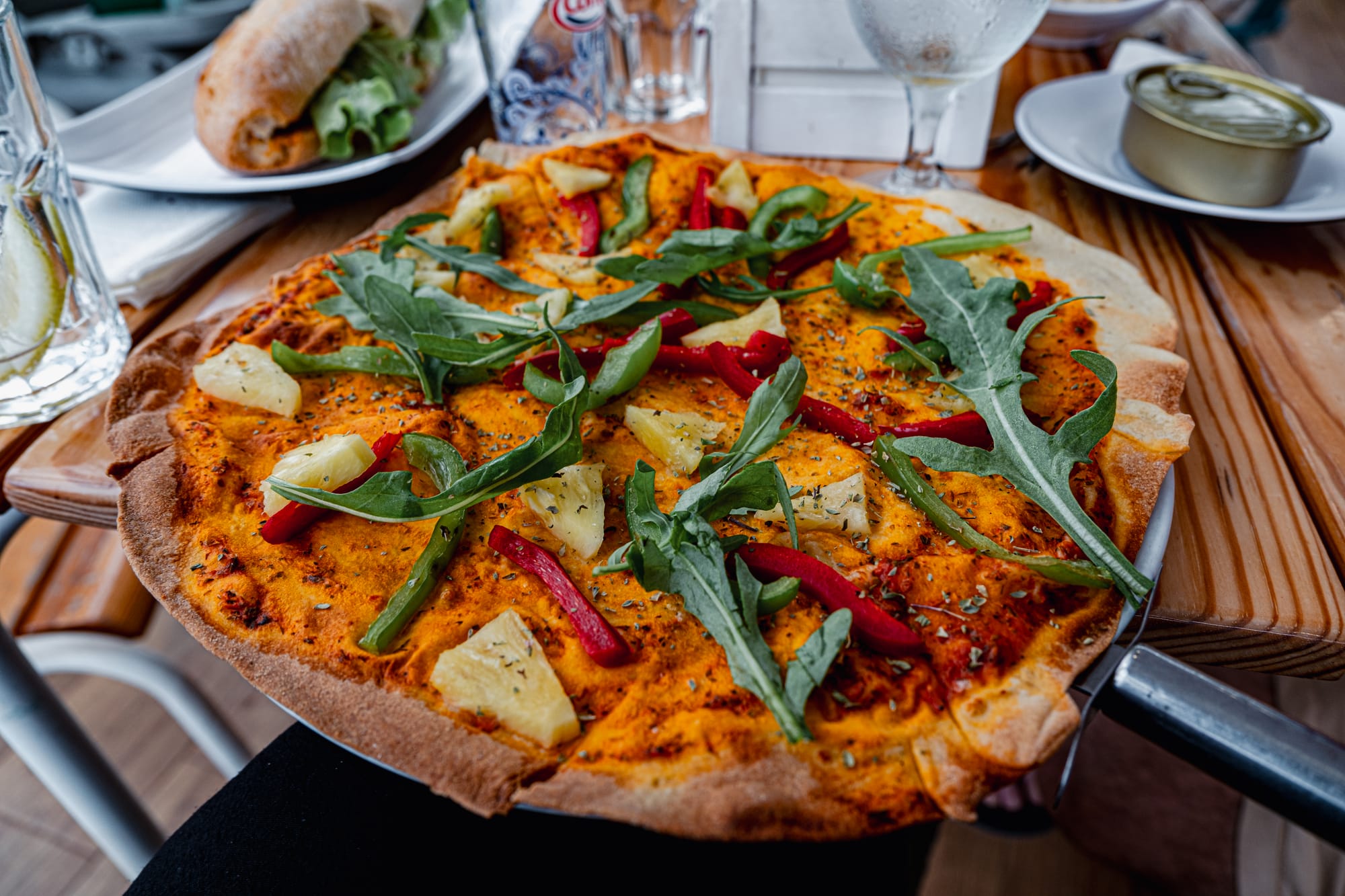 Close-up of a vegan pizza topped with pineapple, bell peppers, and arugula on a wooden table at a beachside restaurant in Ribeira Quente