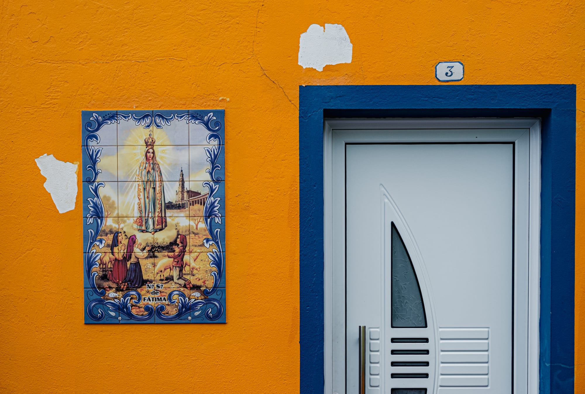 A white door framed in blue on a bold orange wall, with a religious azulejo tile of Nossa Senhora de Fátima beside it in Ribeira Quente