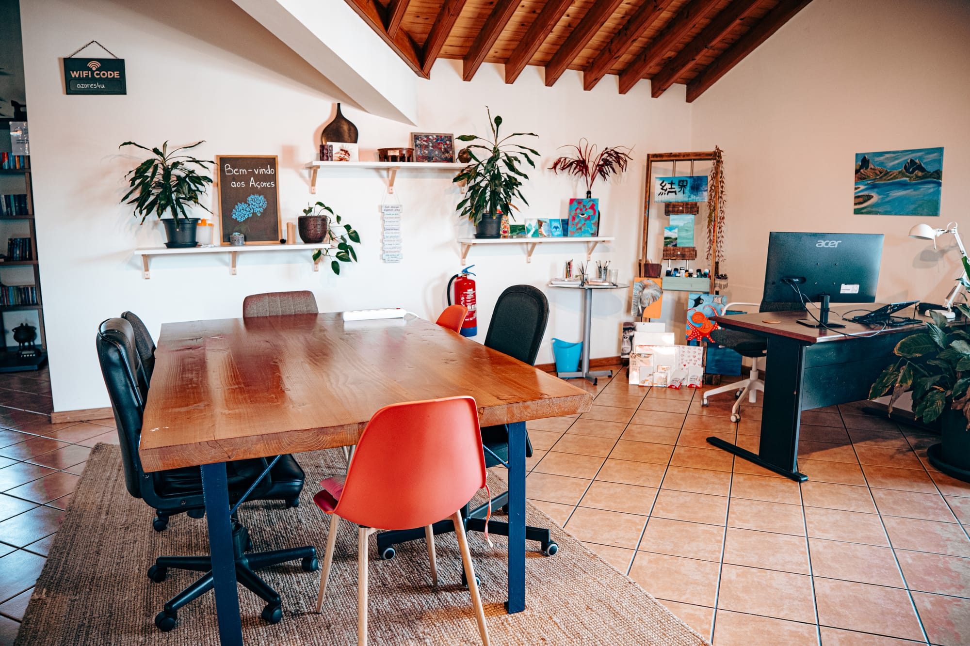Cozy coworking space at Lava Coliving in São Miguel, featuring a large wooden table, colorful chairs, houseplants, and wall art under a vaulted wooden ceiling—blending work-friendly design with island warmth
