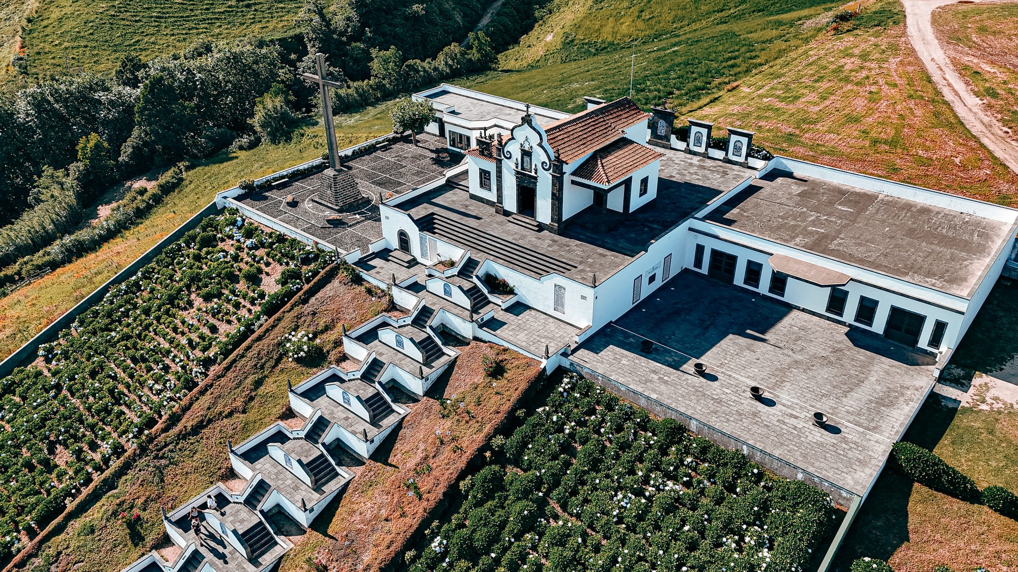 Drone shot of the Santuário de Nossa Senhora da Paz showing the white church, symmetrical staircase with blue tile panels, and surrounding green hills and hydrangea gardens