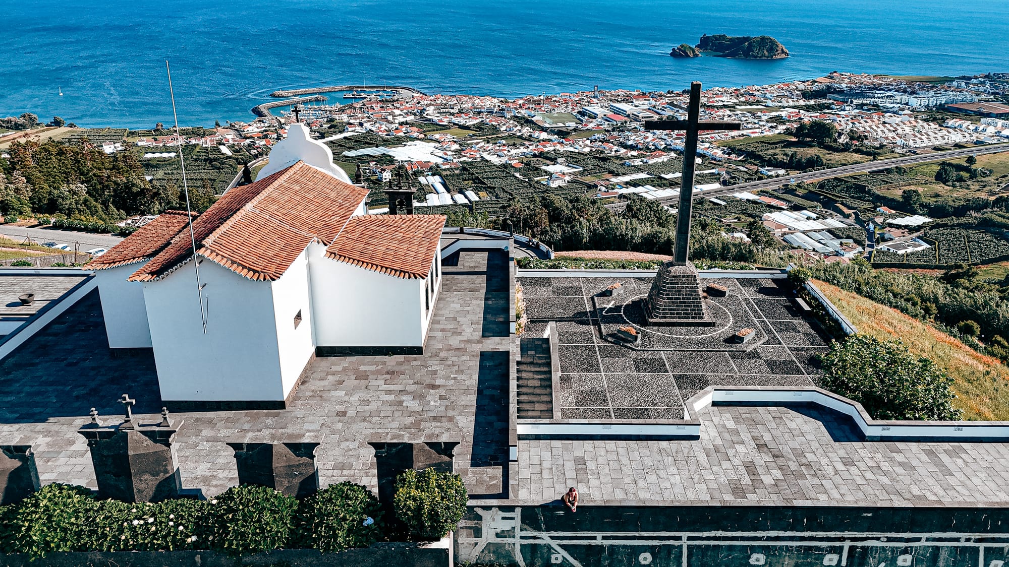 Aerial view from behind the chapel at Santuário de Nossa Senhora da Paz, showing the large stone cross and panoramic backdrop of Vila Franca do Campo and the Atlantic Ocean