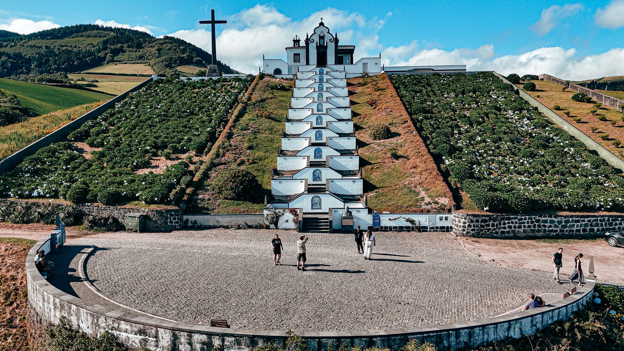 Frontal view of the Santuário de Nossa Senhora da Paz showing the long staircase flanked by flower beds, leading up to the whitewashed chapel with a basalt cross standing to its left