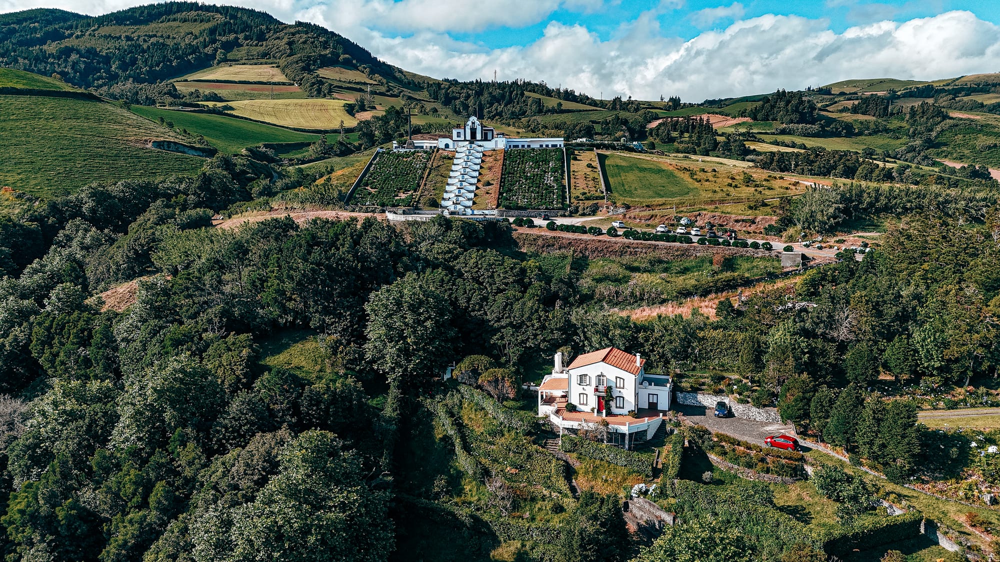 Drone shot of the Santuário de Nossa Senhora da Paz showing its symmetrical staircase ascending the hillside, surrounded by lush green fields, forest, and terraced homes below