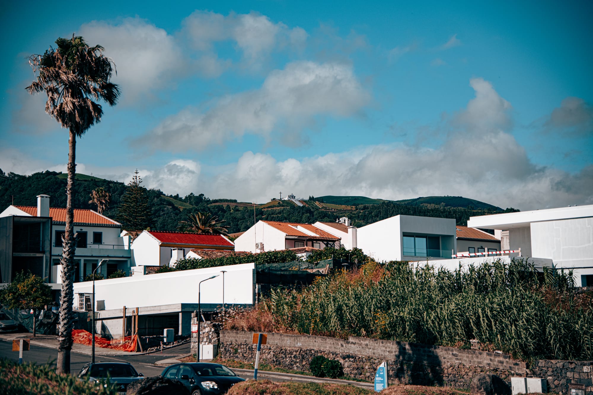 A distant view of Santuário de Nossa Senhora da Paz perched on a hillside above modern houses in Vila Franca do Campo, with a large cross silhouetted against the sky
