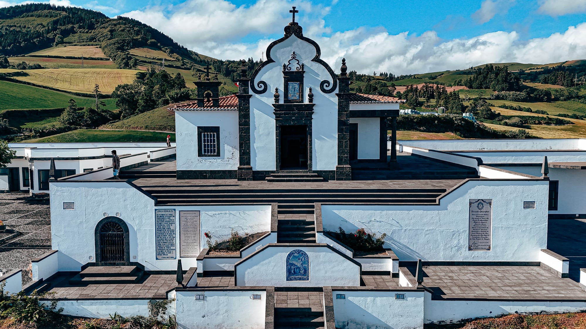 Front-facing view of the whitewashed Santuário de Nossa Senhora da Paz with vivid blue azulejo tiles embedded into the staircase, each depicting biblical scenes, against a lush green hillside backdrop