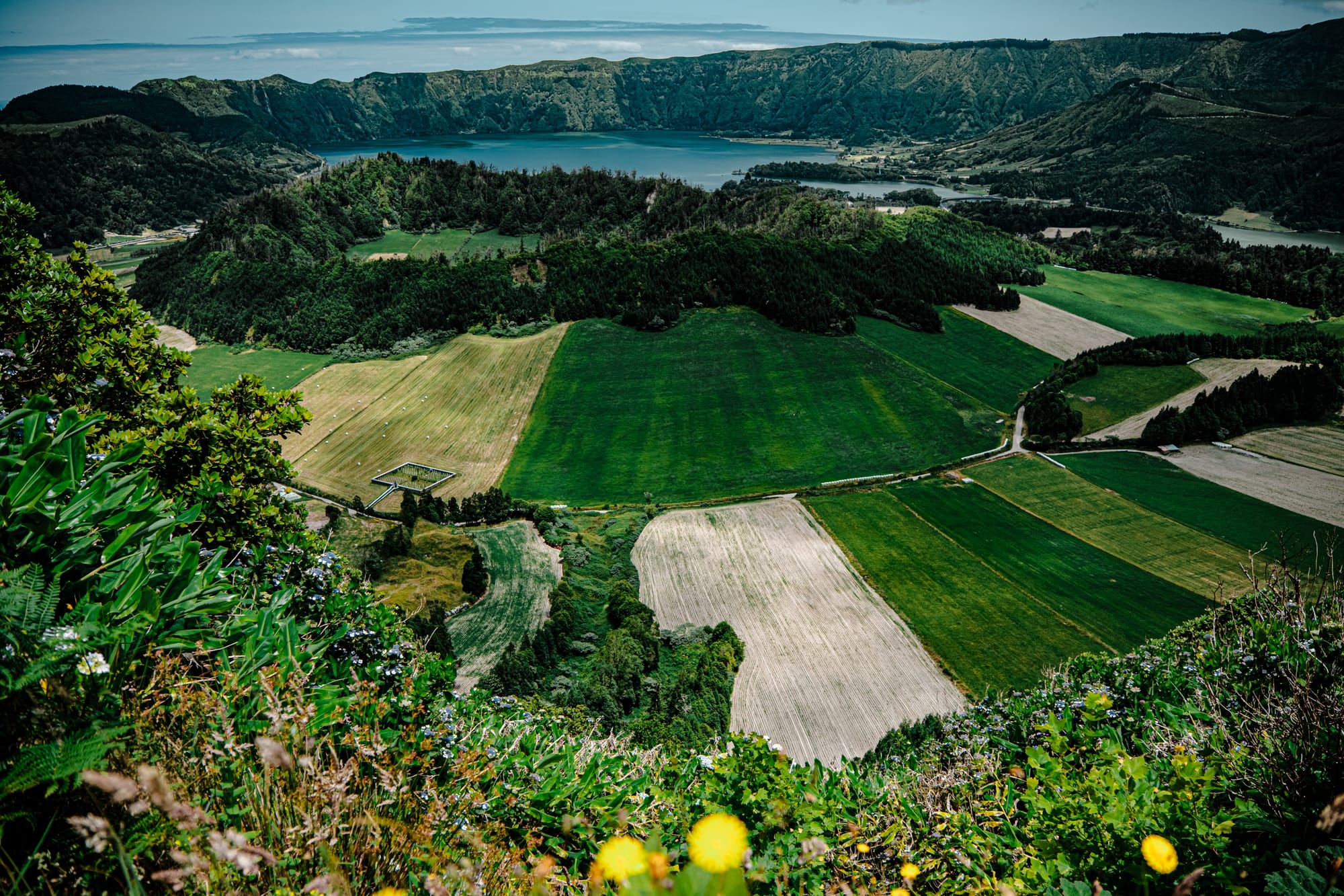 View of green patchwork fields seen from a high vantage point along the PR3-SMI trail, with Lake Sete Cidades and surrounding volcanic ridge in the distance