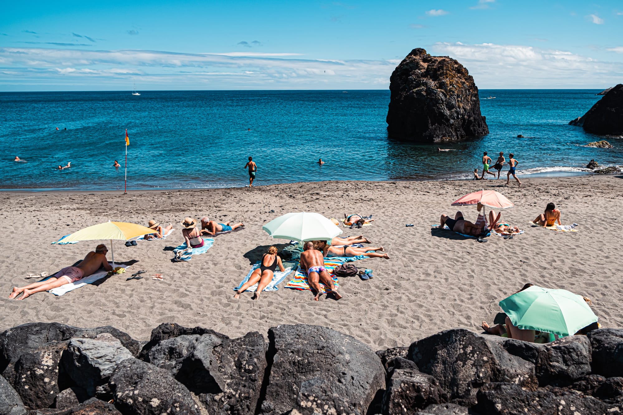 View of sunbathers on Praia Vinha d’Areia beach with umbrellas and swimmers near volcanic rock formations in the sea