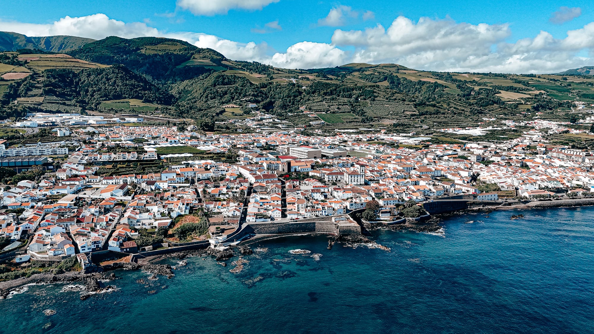 Drone shot showing the red rooftops and coastline of Vila Franca do Campo, with green hills rising behind the town