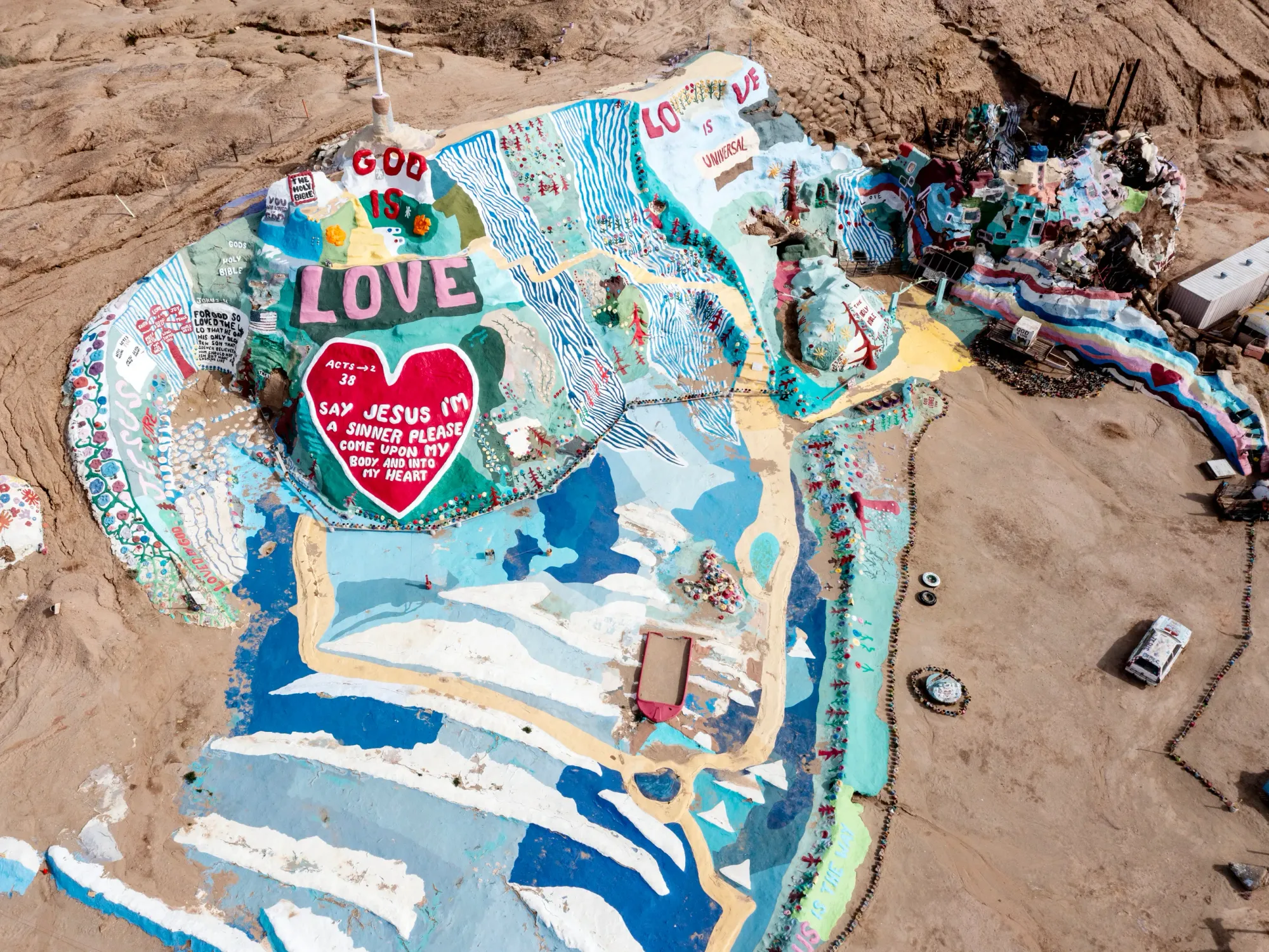 An aerial view of Salvation Mountain in the California desert, a brightly painted man-made hill with colorful stripes, murals, and large text reading “God is Love"