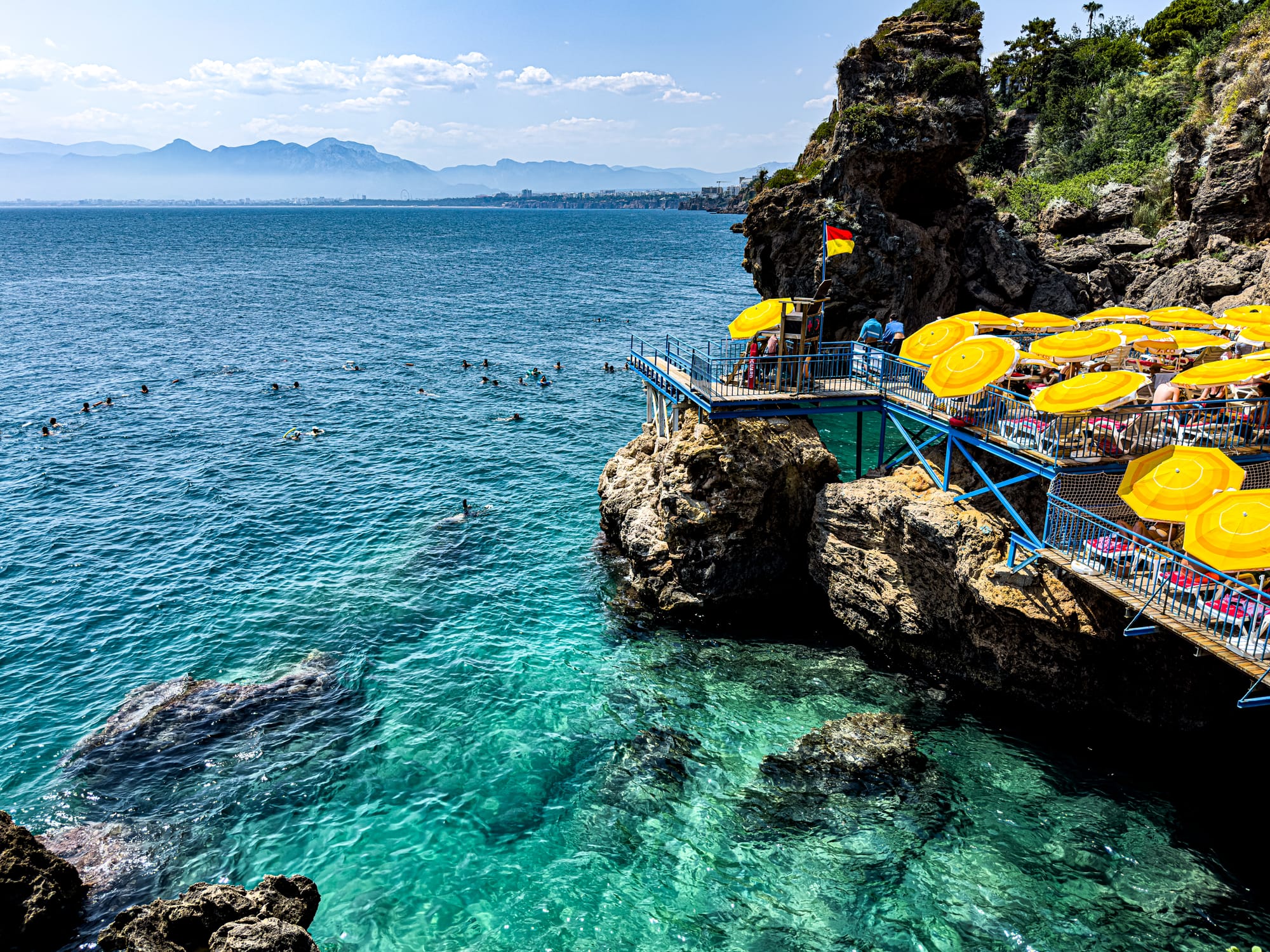 Swimmers floating in clear turquoise water beside the rocky platforms and yellow umbrellas of İnciraltı Beach in Antalya, Turkey