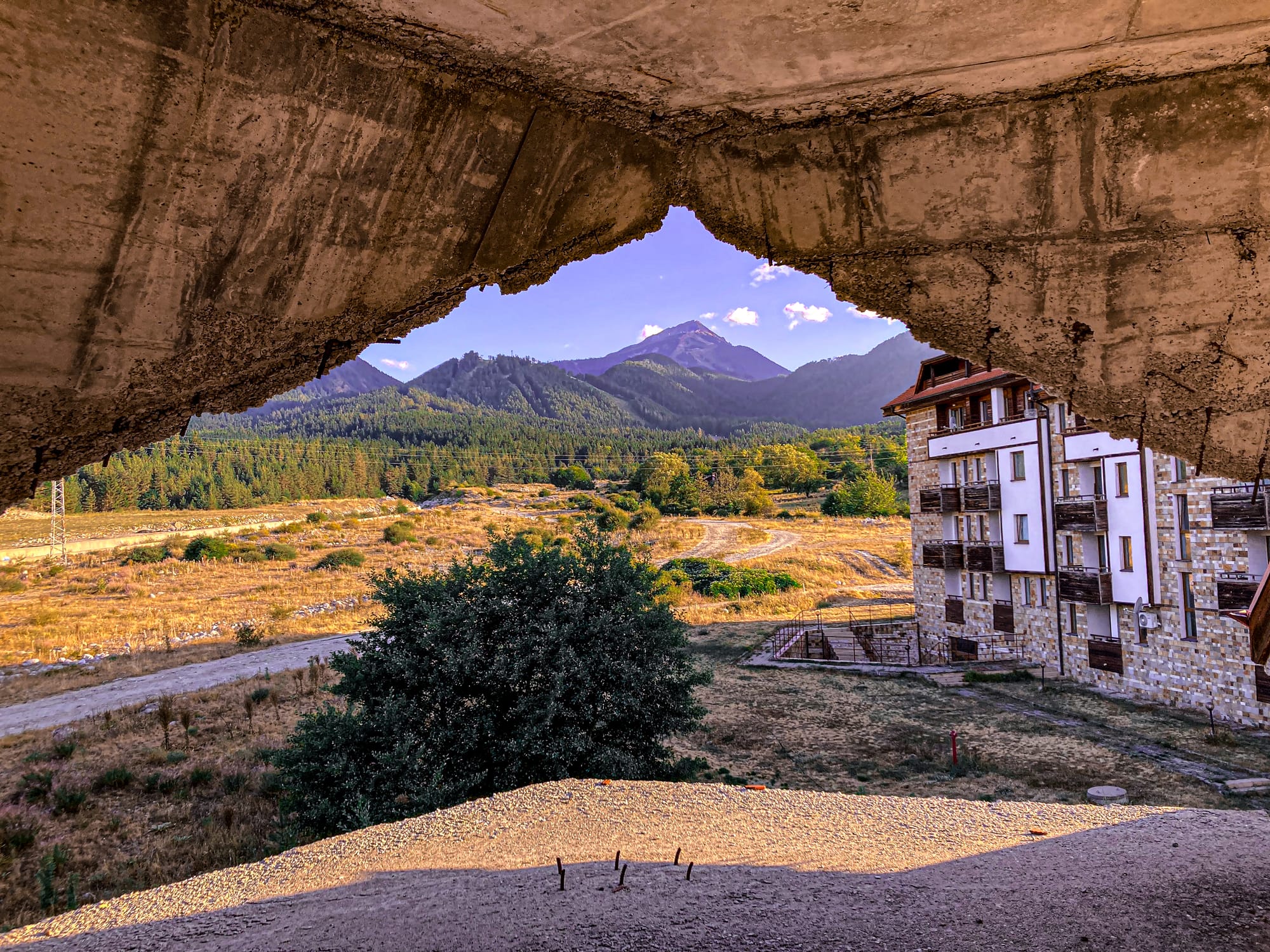 Landscape of the Pirin mountains seen through a jagged concrete opening of an unfinished building in Bansko, Bulgaria, with a modern apartment block and dry field below