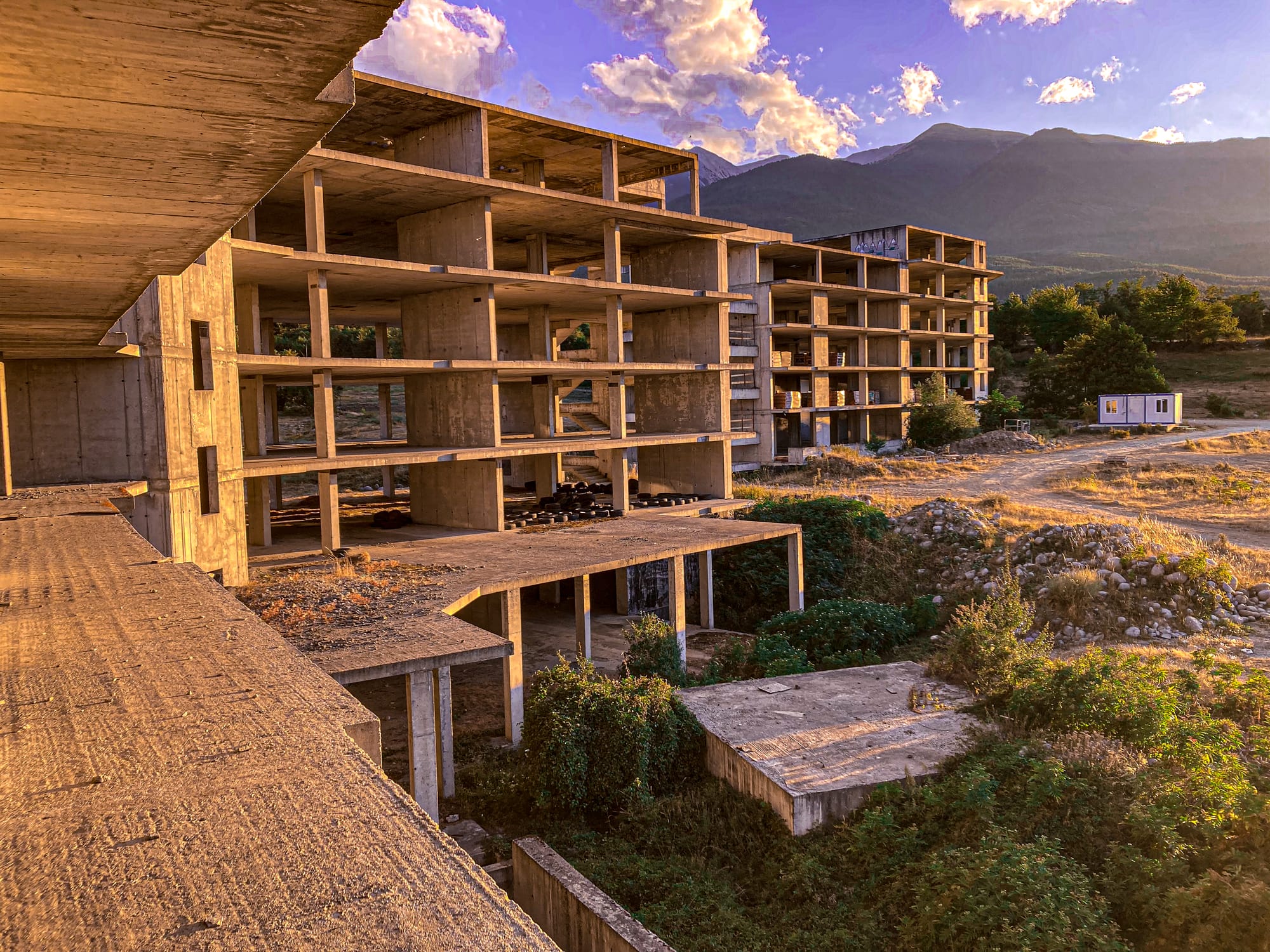 Unfinished concrete apartment blocks in Bansko, Bulgaria, bathed in evening sunlight, with the Pirin mountains in the background and overgrown vegetation around the base