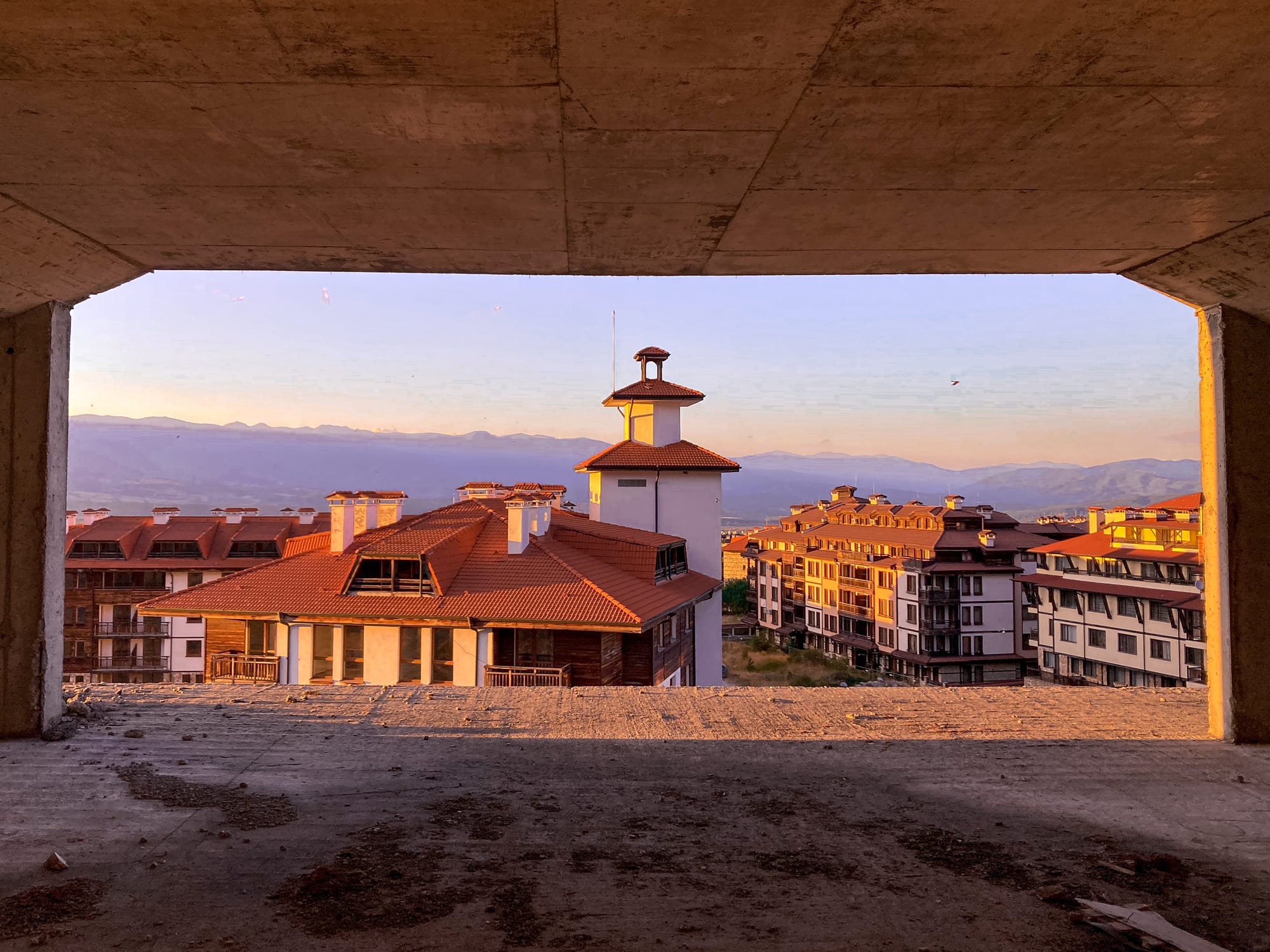 Looking out from the inside of an unfinished concrete building in Bansko, Bulgaria, toward red-roofed apartments and a tower in warm sunset light, with mountains visible in the distance