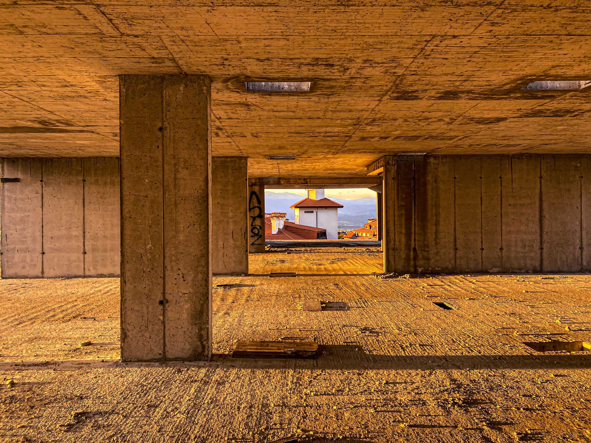 Interior of an unfinished concrete building in Bansko, Bulgaria, with a square opening framing red-tiled rooftops and hills beyond, lit by warm evening sunlight