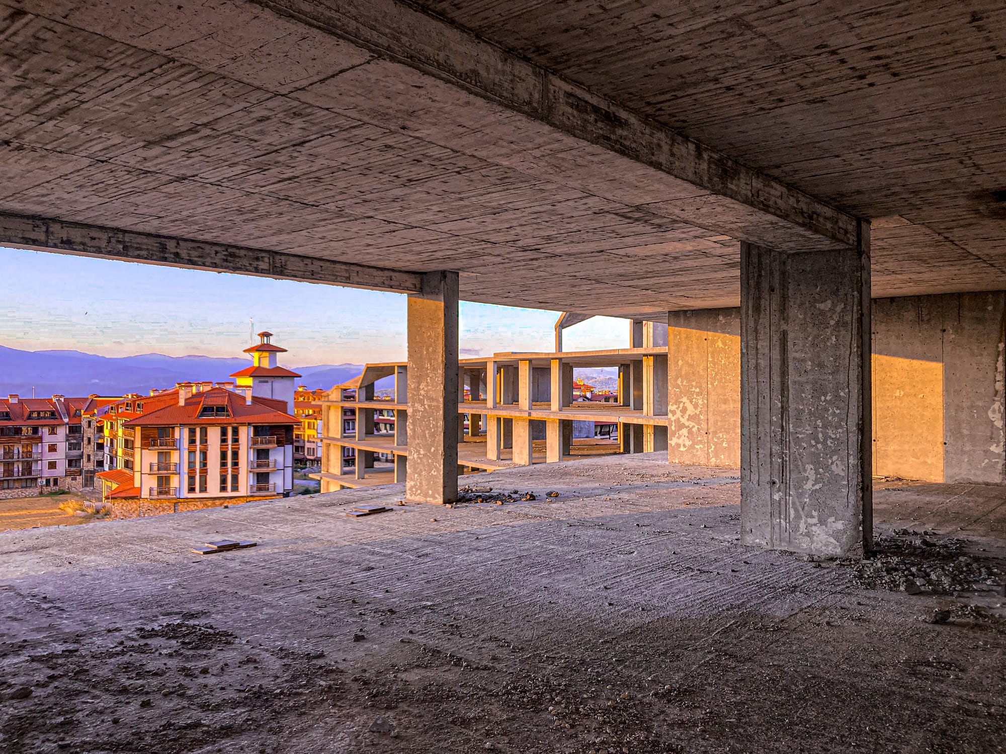 Interior of an unfinished concrete building in Bansko, Bulgaria, with views of completed apartment blocks and rooftops outside, bathed in warm evening light
