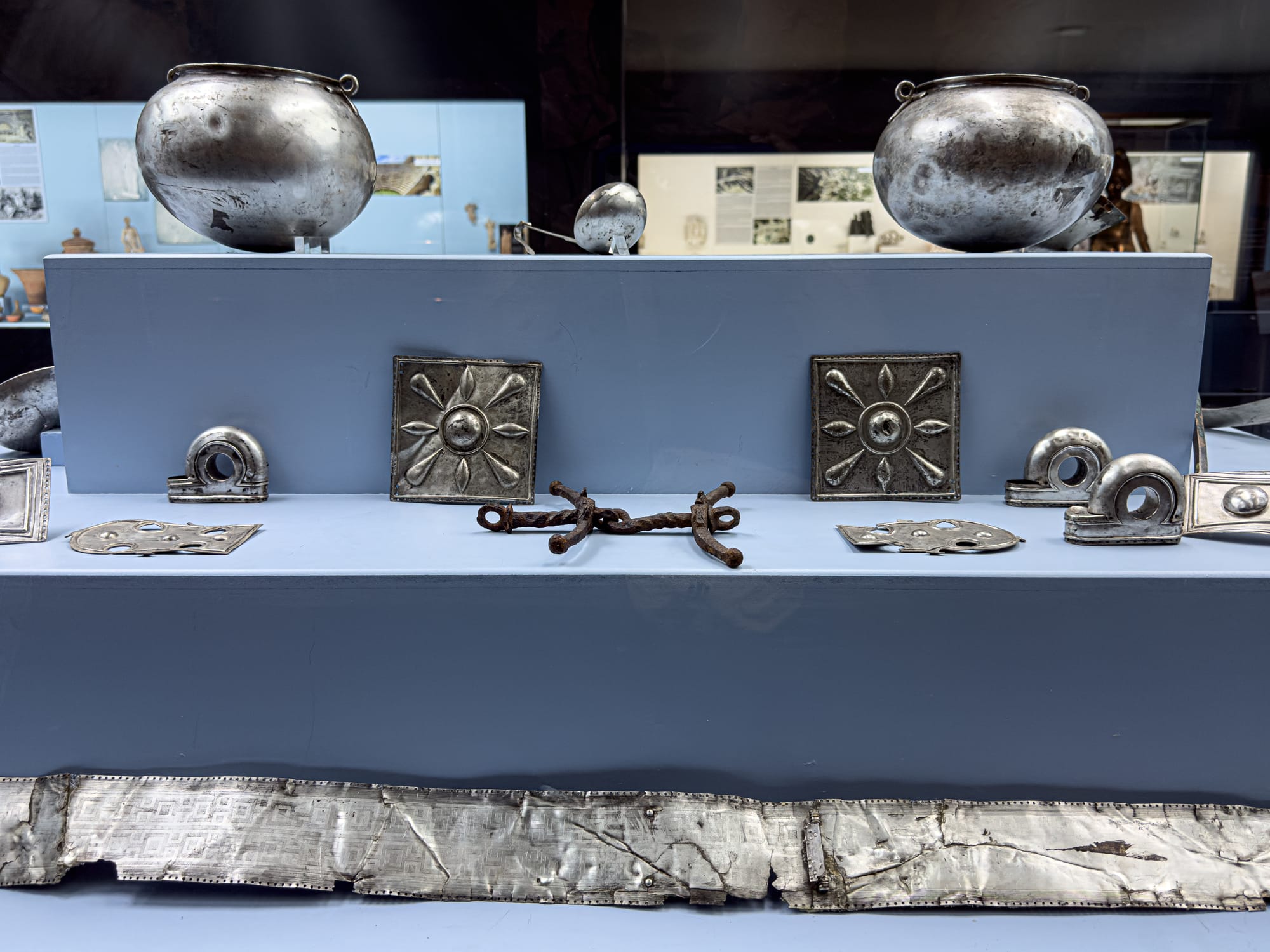 Display of ancient silver artifacts including pots, floral-patterned plaques, and decorative metalwork arranged on a soft blue platform in Antalya’s Archaeological Museum