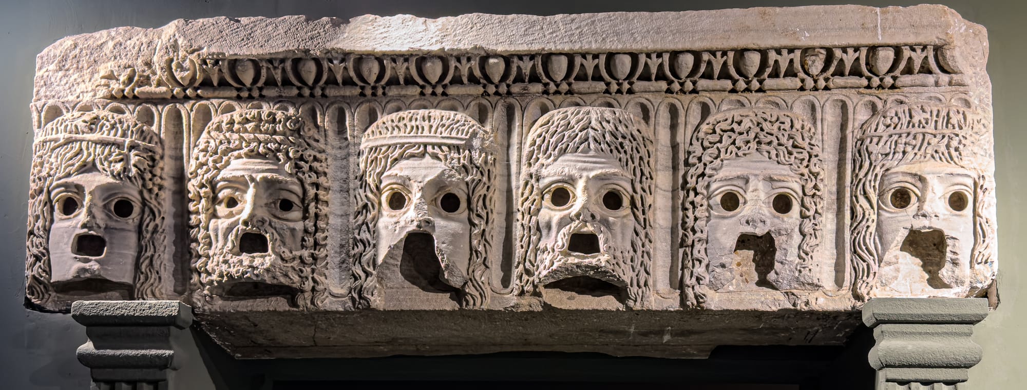 Stone carving of six theatrical masks with wide eyes and open mouths, lined up in dramatic expression