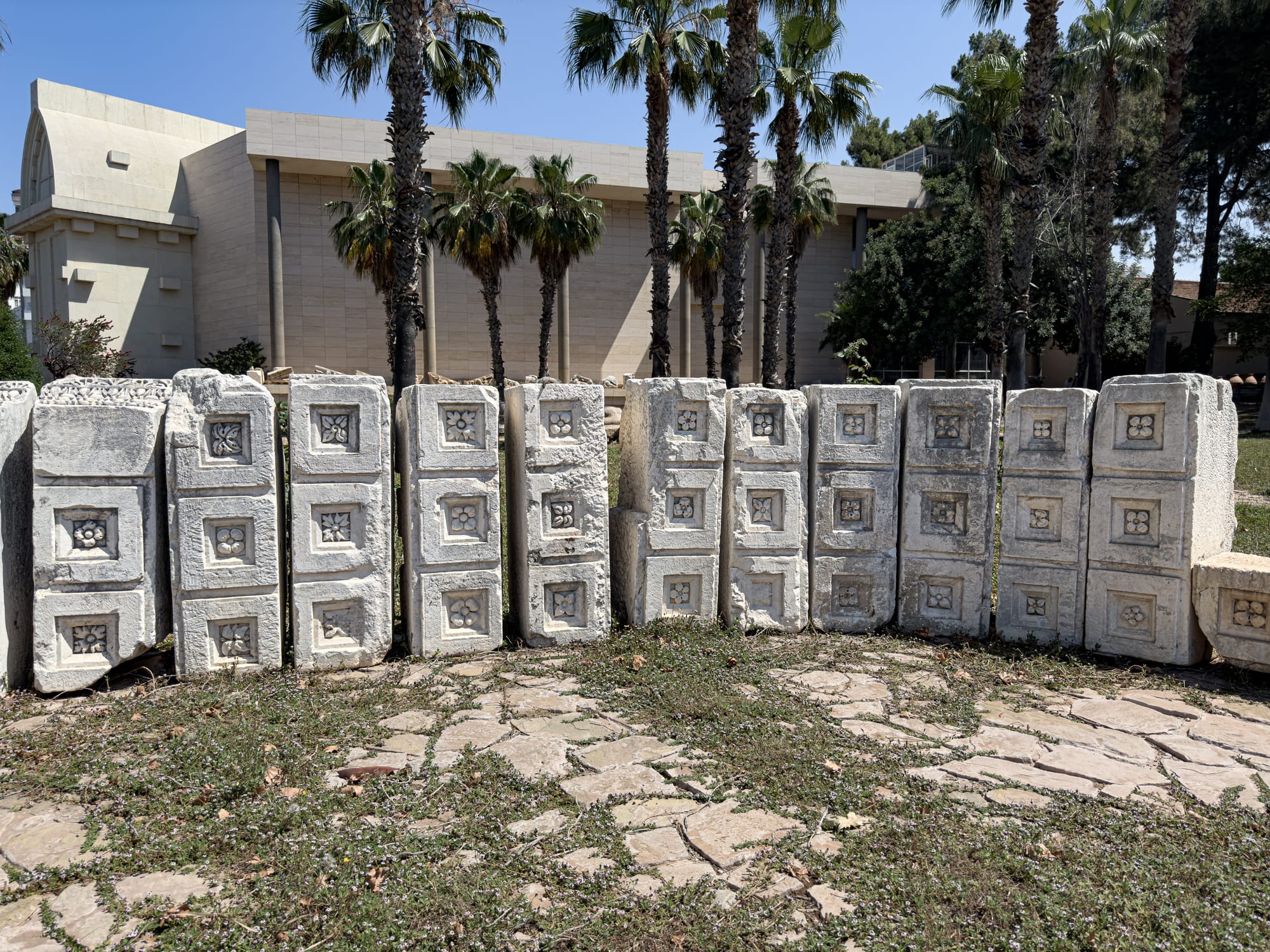 Stone blocks with carved square and flower patterns arranged in a line on the lawn outside Antalya’s Archaeological Museum, framed by palm trees and the museum building