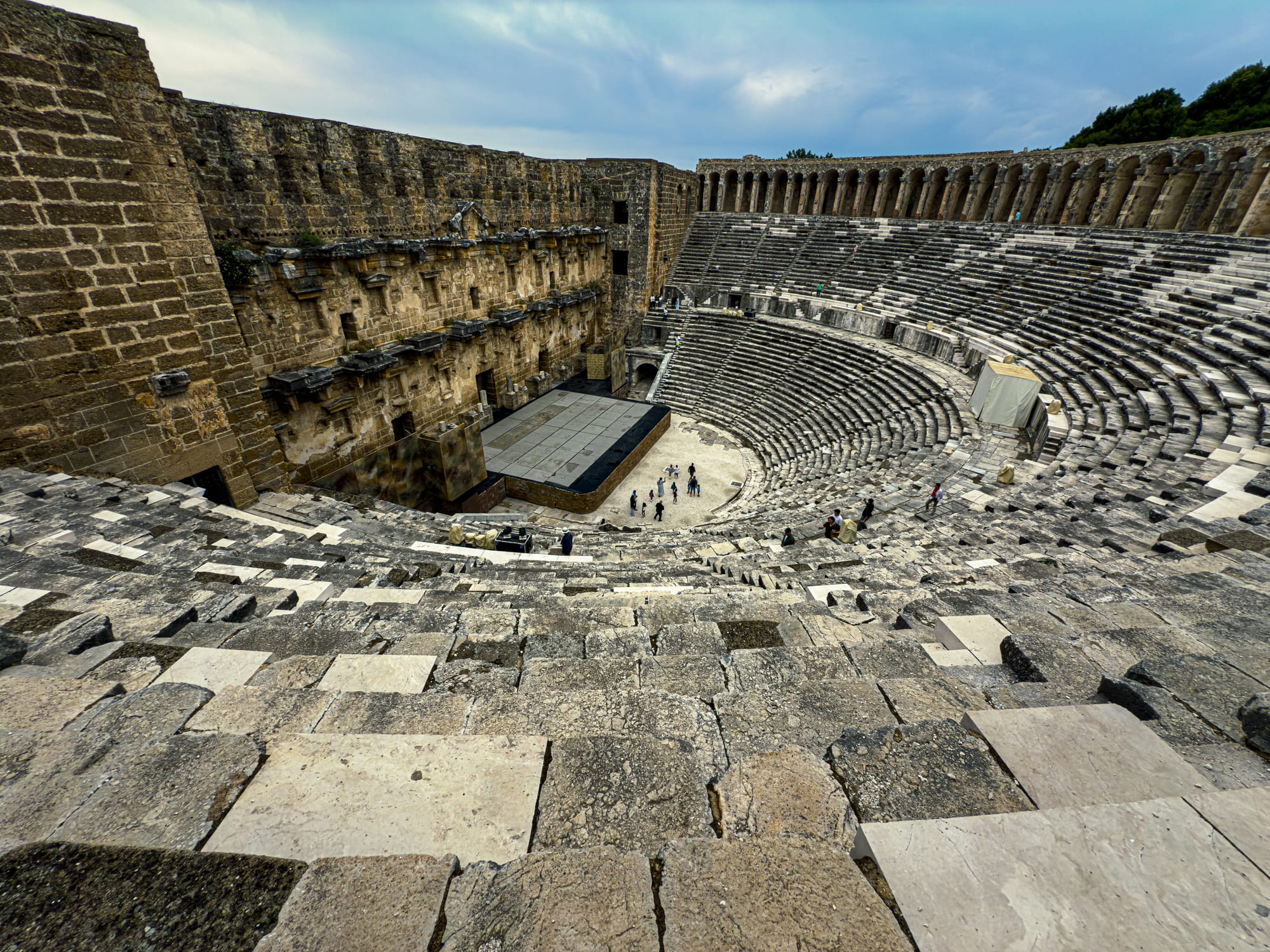 A wide-angle view of the Aspendos Roman theater shows its vast semicircular seating and preserved stage structure under a cloudy sky