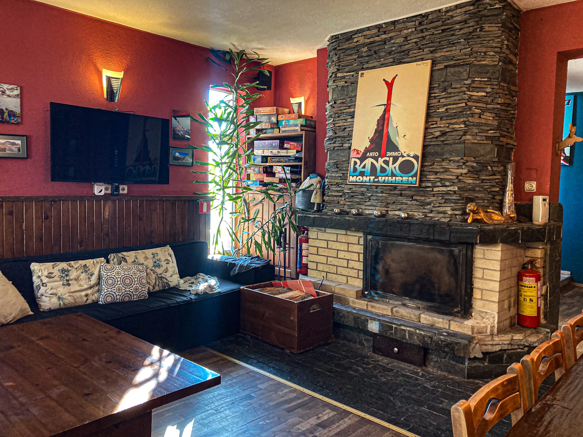 Cozy communal lounge at Avalon Coliving in Bansko, featuring a stone fireplace with a vintage Bansko ski poster above it, a black sofa with patterned cushions, a TV on the wall, and a stack of board games in the corner