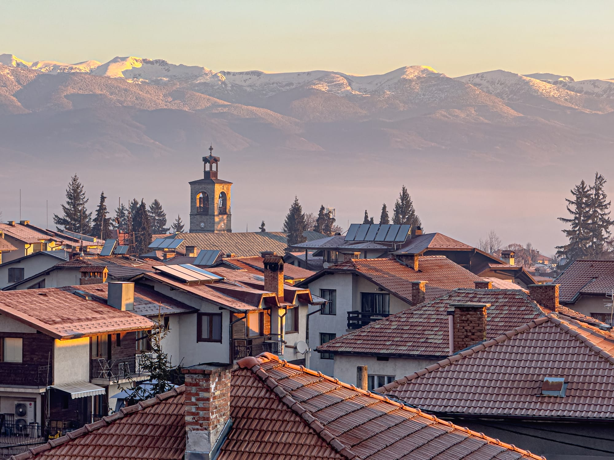 View from a top-floor room at Avalon Coliving in Bansko, looking out over terracotta rooftops, the town’s church tower, and the snow-capped Pirin mountains glowing in the soft light of sunrise
