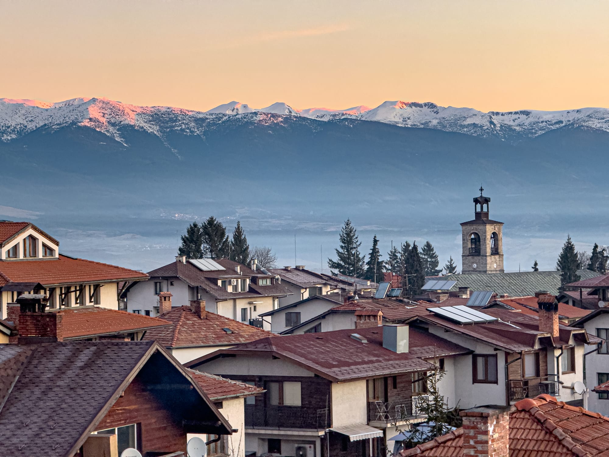 View from a bedroom window at Avalon Coliving in Bansko, showing red-tiled rooftops, the town’s church tower, and the Pirin mountains in the background with their peaks dusted in snow under a soft orange sunrise