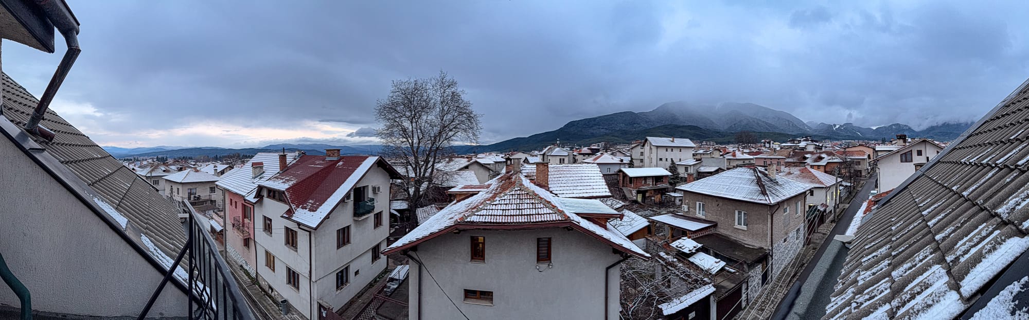Panoramic view from a top-floor balcony at Avalon Coliving in Bansko, overlooking snow-dusted rooftops of the town with the Pirin mountains in the distance under a cloudy winter sky
