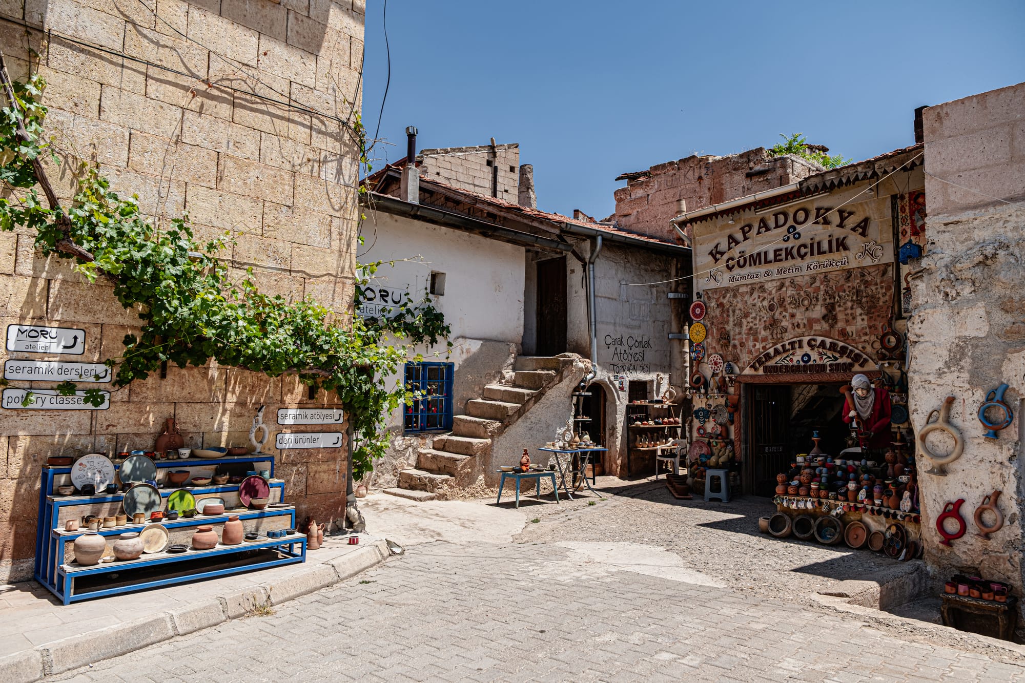 Outdoor view of pottery shops and studios in Avanos, Cappadocia, with shelves displaying colorful ceramic plates, bowls, and clay pots under bright sunlight, surrounded by stone buildings and green vines